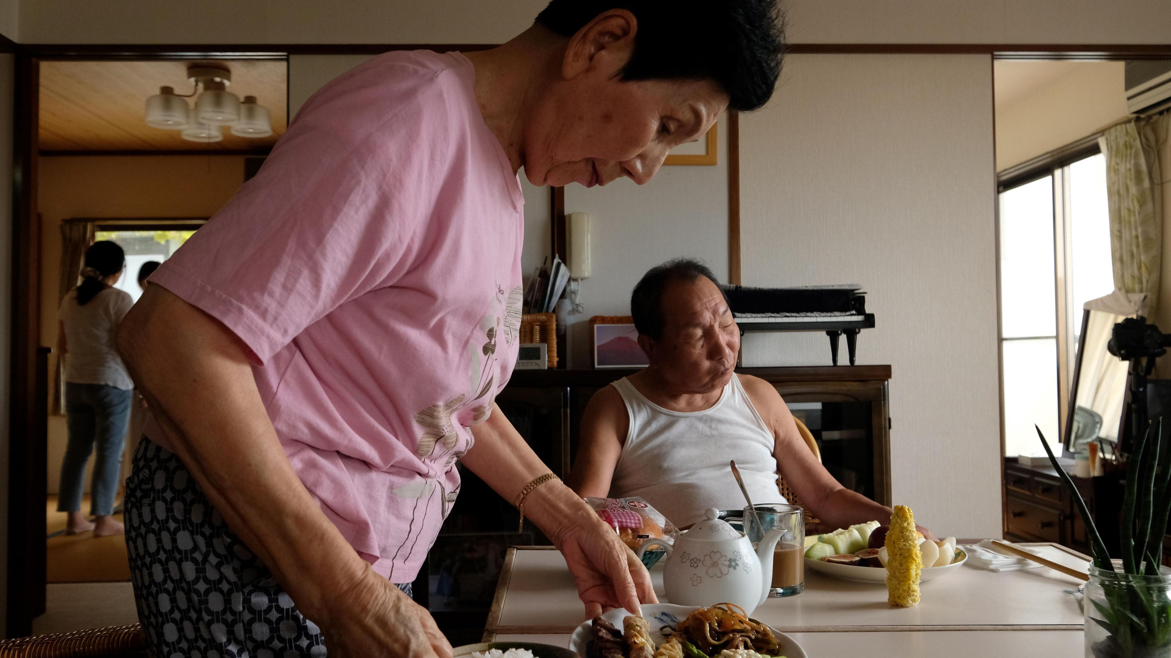 A woman in a pink shirt puts meals down on a kitchen table while a man in a white singlet sits at the head of the table