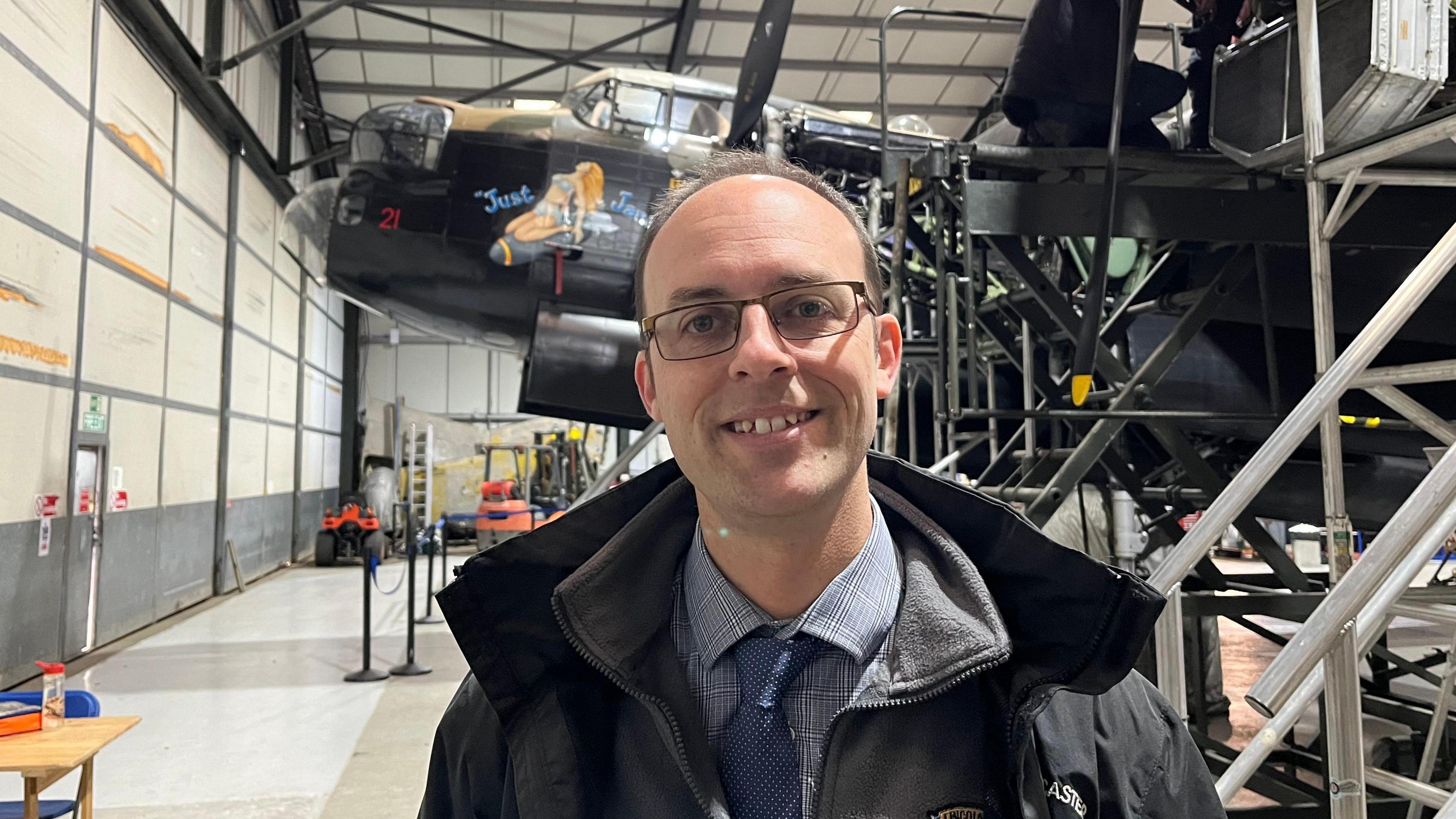 A head and shoulders shot of Andrew Panton smiling at the camera. He has short brown hair and is wearing black glasses. He is also wearing a light blue check shirt, a blue spotty tie, a grey fleece and a black waterproof coat. He is stood in a warehouse with Just Jane behind him. 
