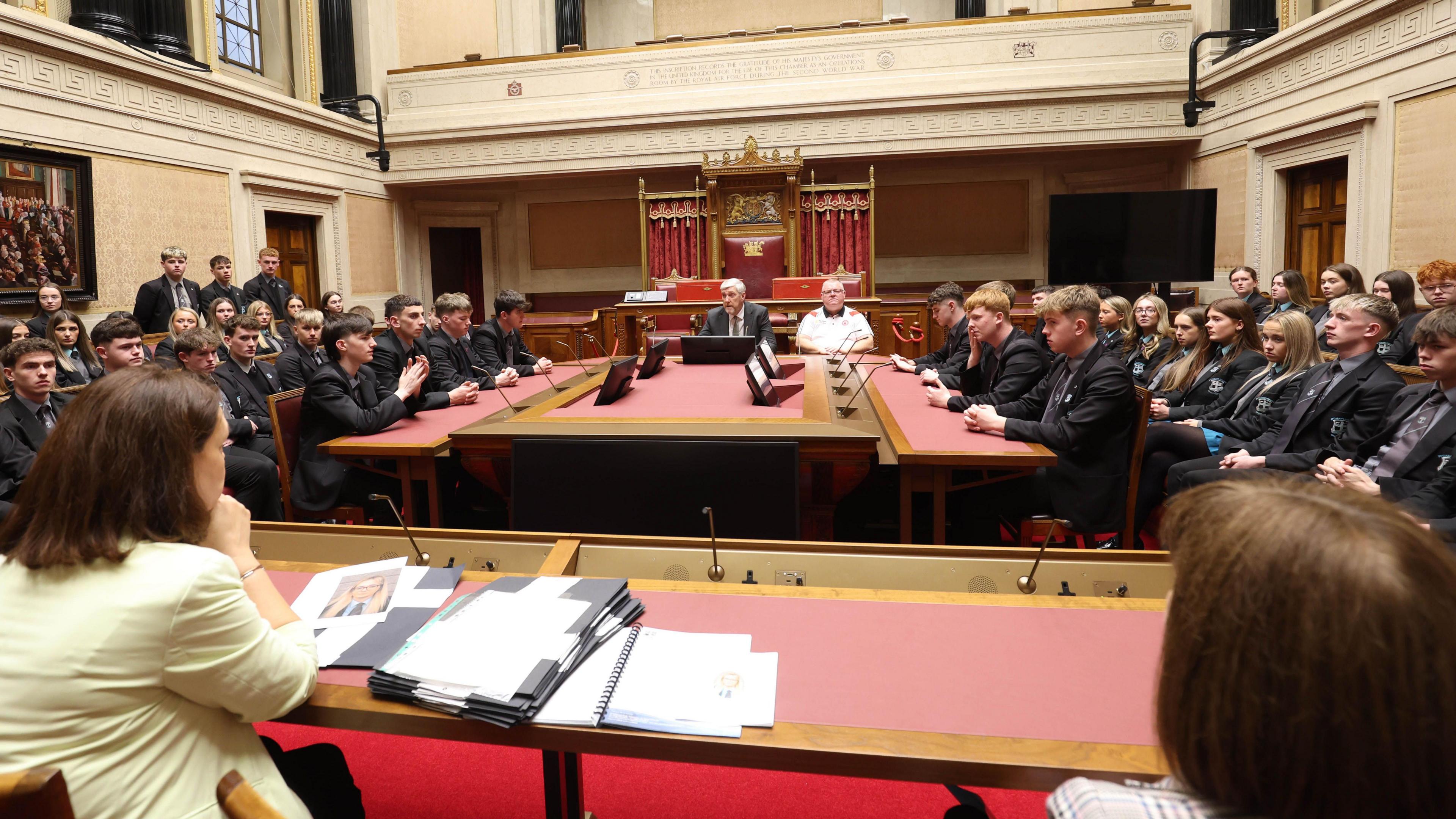 Stormont Senate chamber showing minister O'Dowd in the middle, surrounded by pupils.