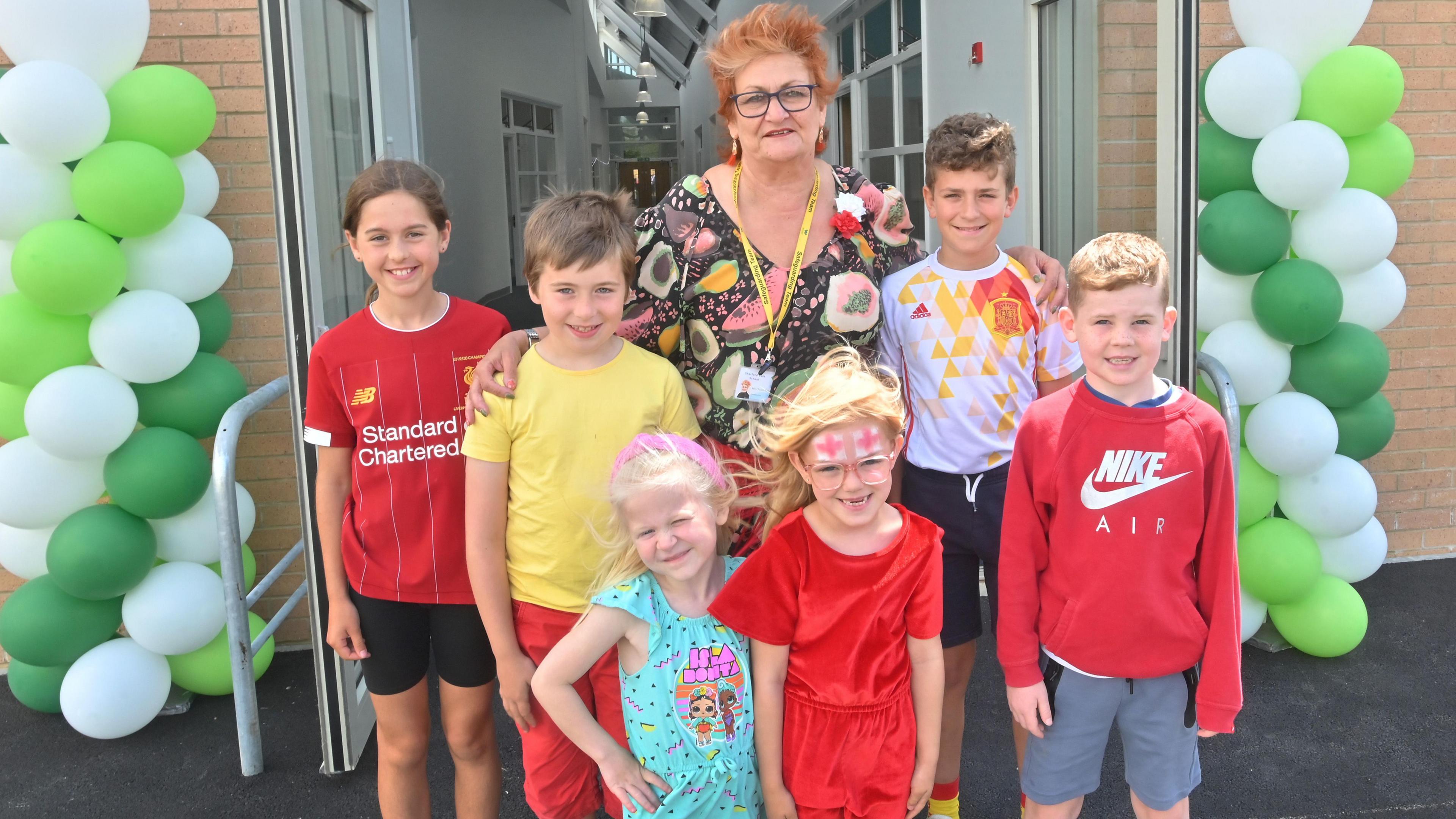 Headteacher, Susie Evans in a brown, cream and pink patterned dress with six pupils from the school standing in front of a balloon arch at the entrance to the new wing