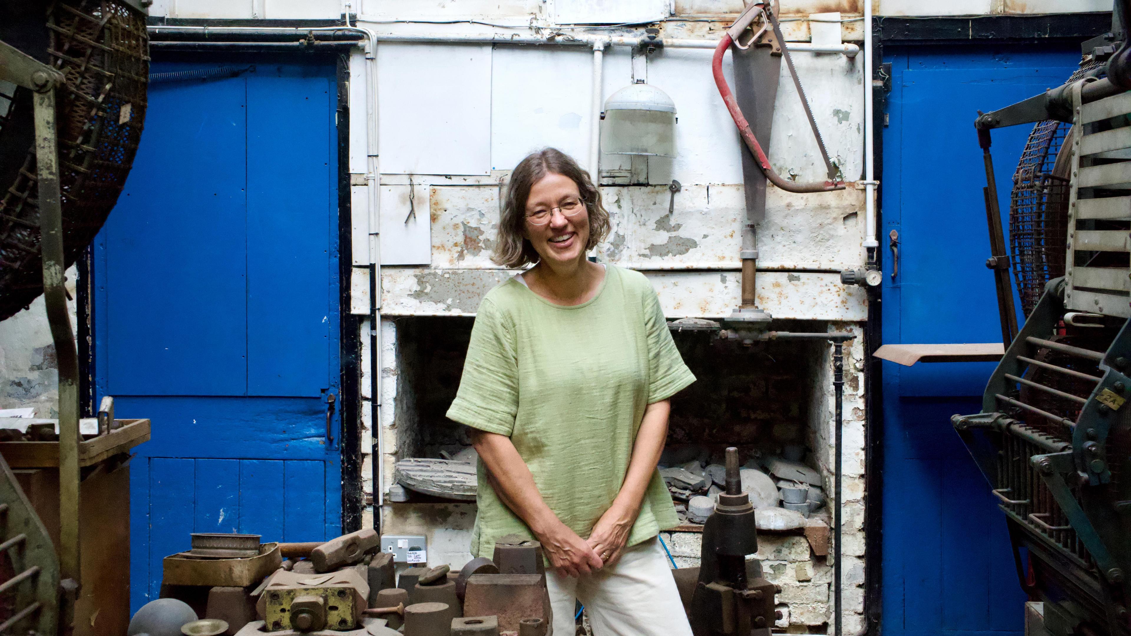 A woman with brown hair and glasses in a green top stood in front of a white wall and blue door in an old factory