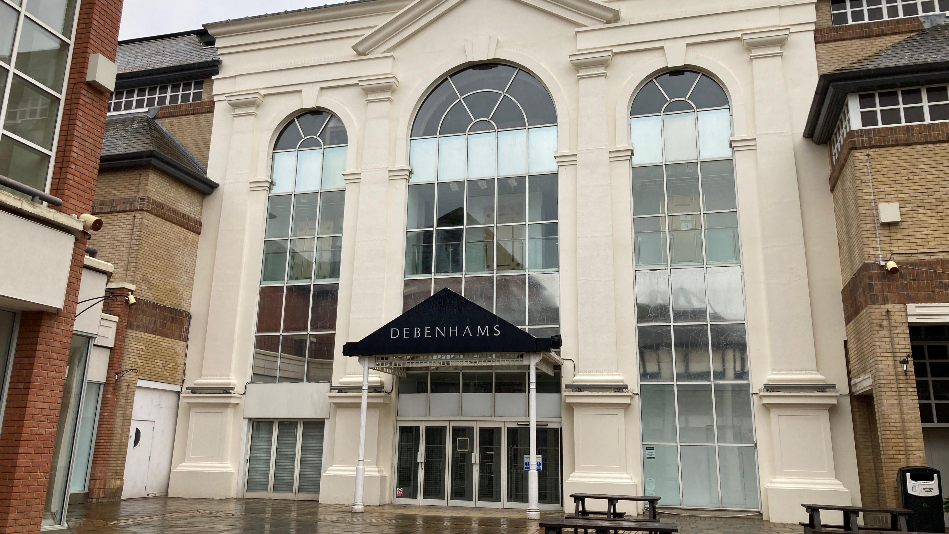 The large, vacant Debenhams building. It is three levels tall and has large, archway-shaped windows split between cream pillars. The building faces on to an outdoor shopping street. It is pictured on a rainy day.