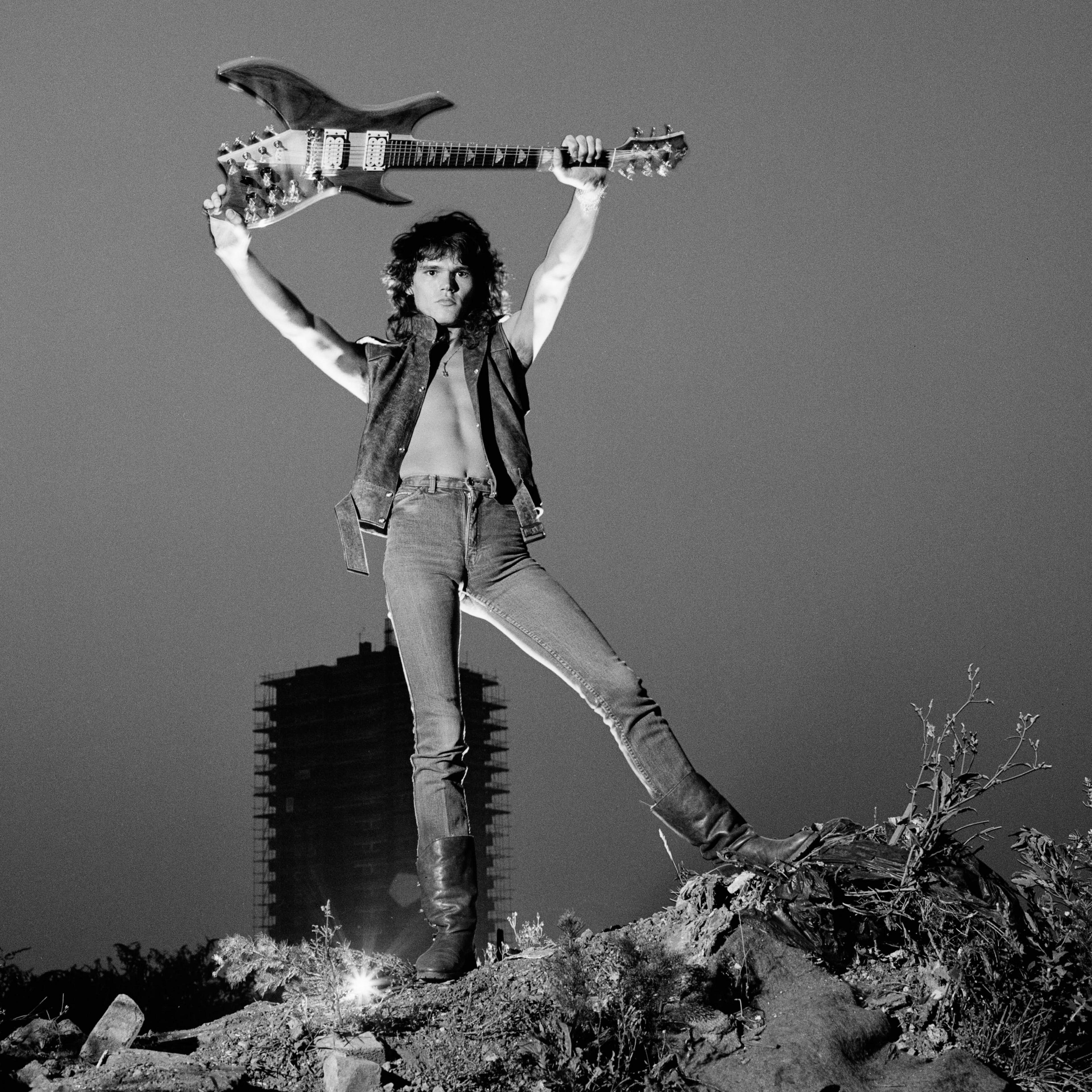 A black and white image of Robin George on top of a rocky outcrop holding his guitar aloft. He is wearing a short-sleeved jacket, jeans and boots and is bare chested.