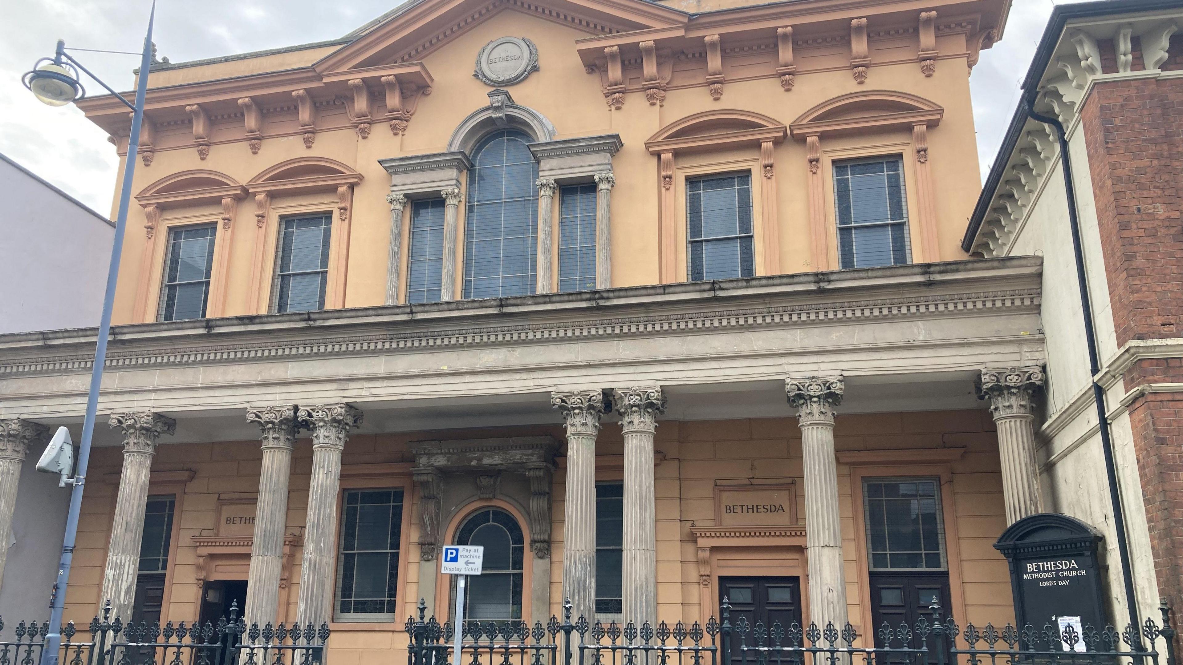 The outside of the chapel has ornamental columns and an arched window on the top floor. There are black metal railings at the front with a road sign and a street light.