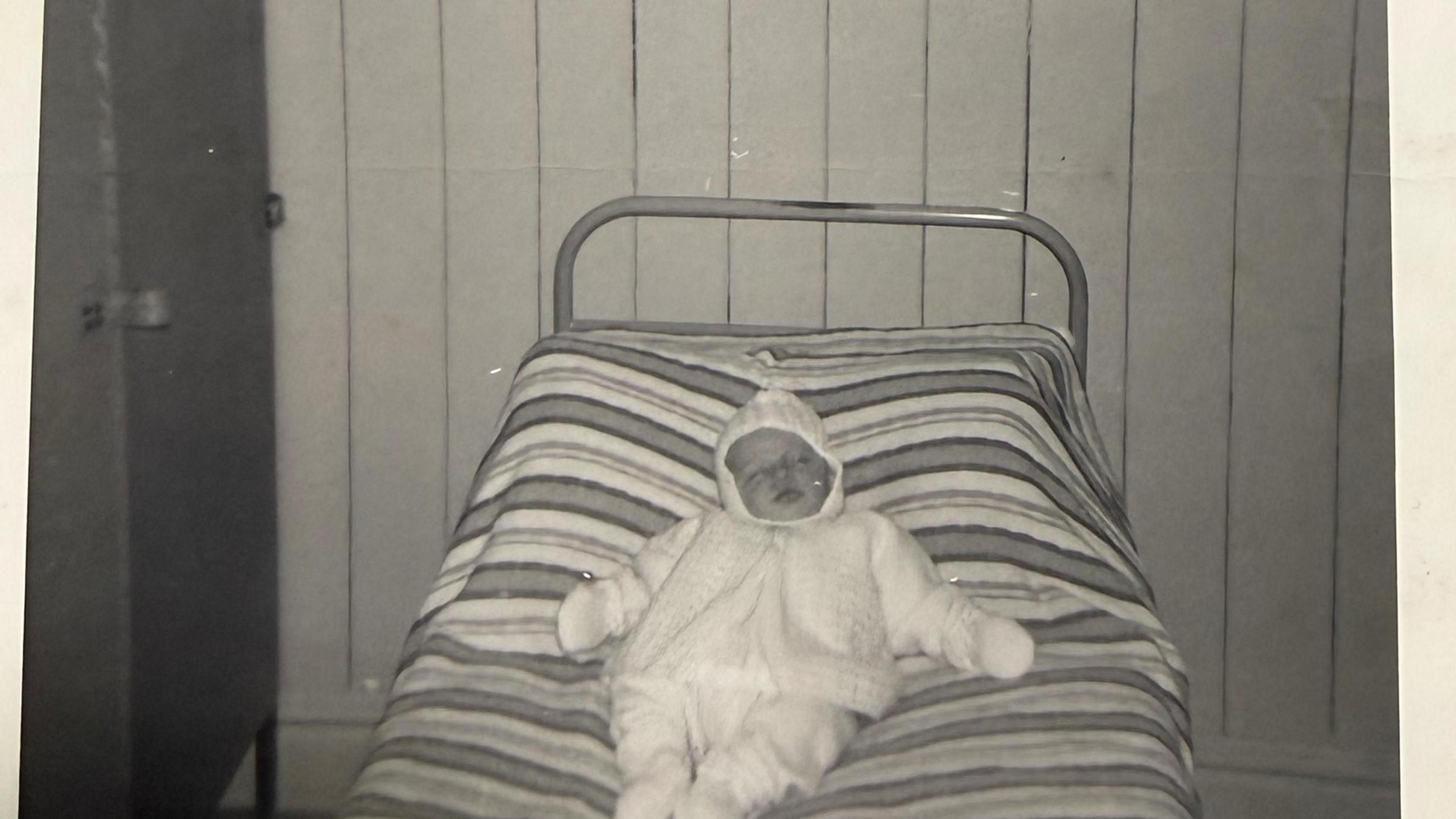 Black-and-white photograph of a small bed with a striped blanket, with a baby Julian laid on top, in a room at St Monica's with wooden wall panels