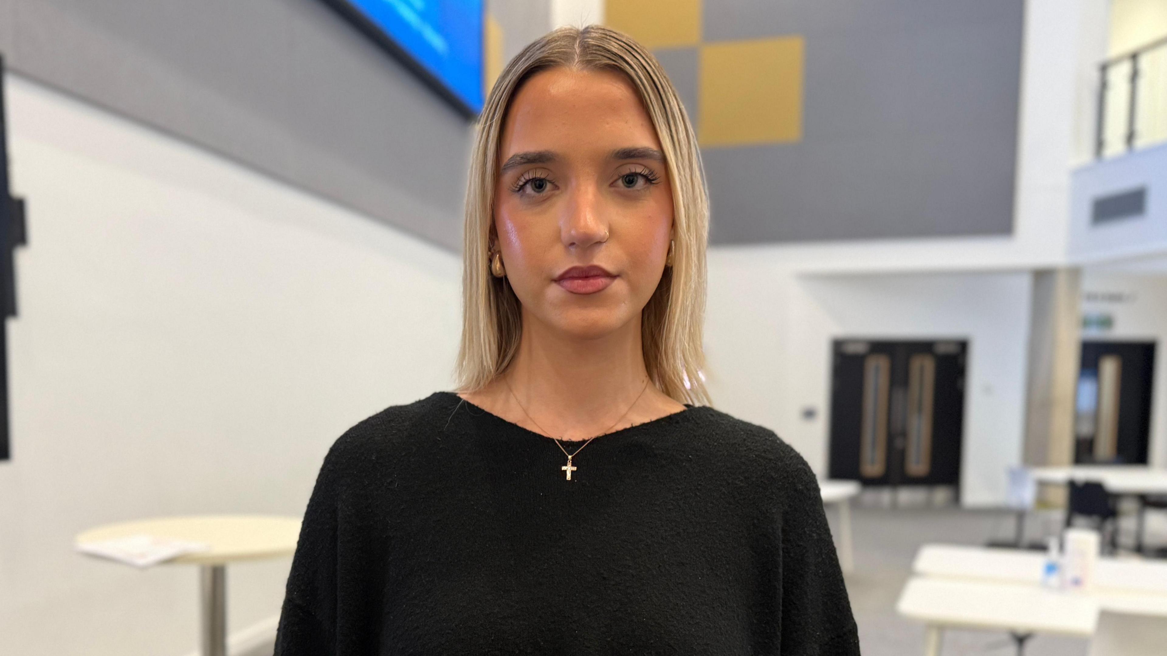 A blonde woman looking directly at the camera wearing a black jumper and a small cross pendant necklace. 