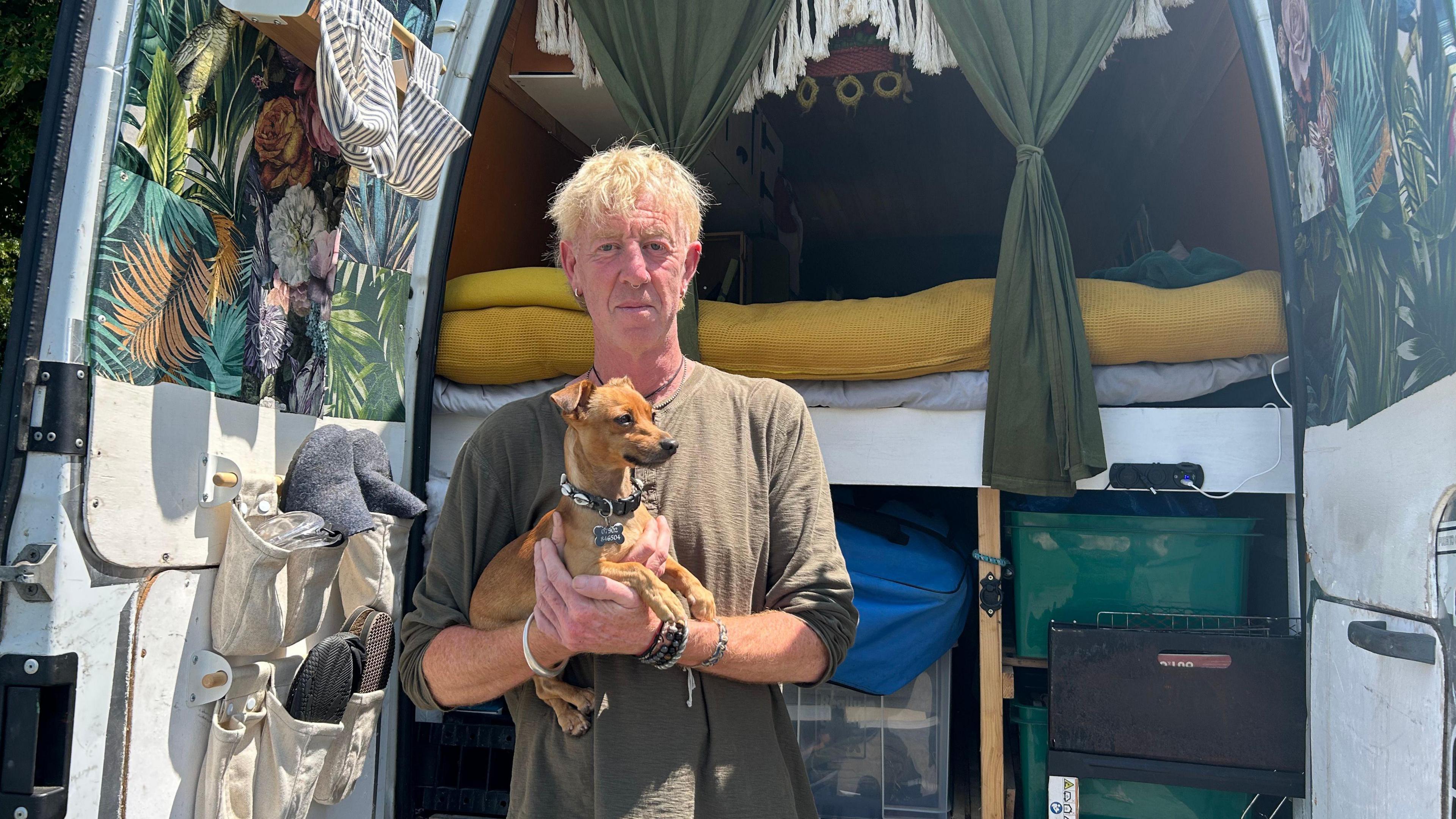 Jasper, who has dyed blonde hair, looks into the camera while holding a small dog. He stands in front of the back of a campervan with the doors open. The van has tasselled green curtains and a bed with a yellow blanket.