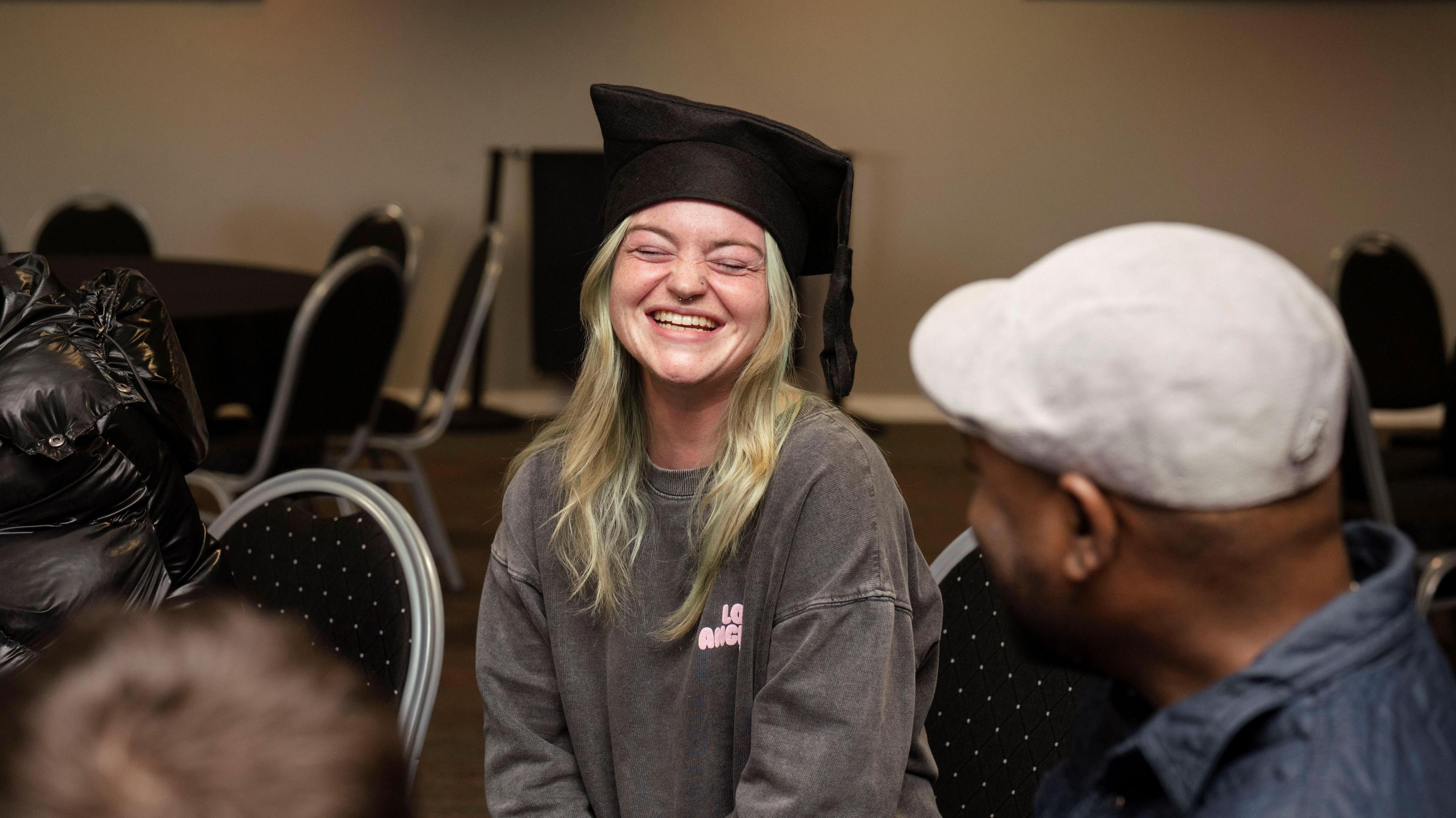 Stephanie is sitting on a  chair with a group of people and laughing. She has long blonde hair and a pink t-shirt on. She is wearing a graduation cap. There are two other people in the photo, you cannot see their face but the back of their heads. The guy on the right has a grey cap and blue shirt. And the one on the left, you can just see his dark brown hair. 
