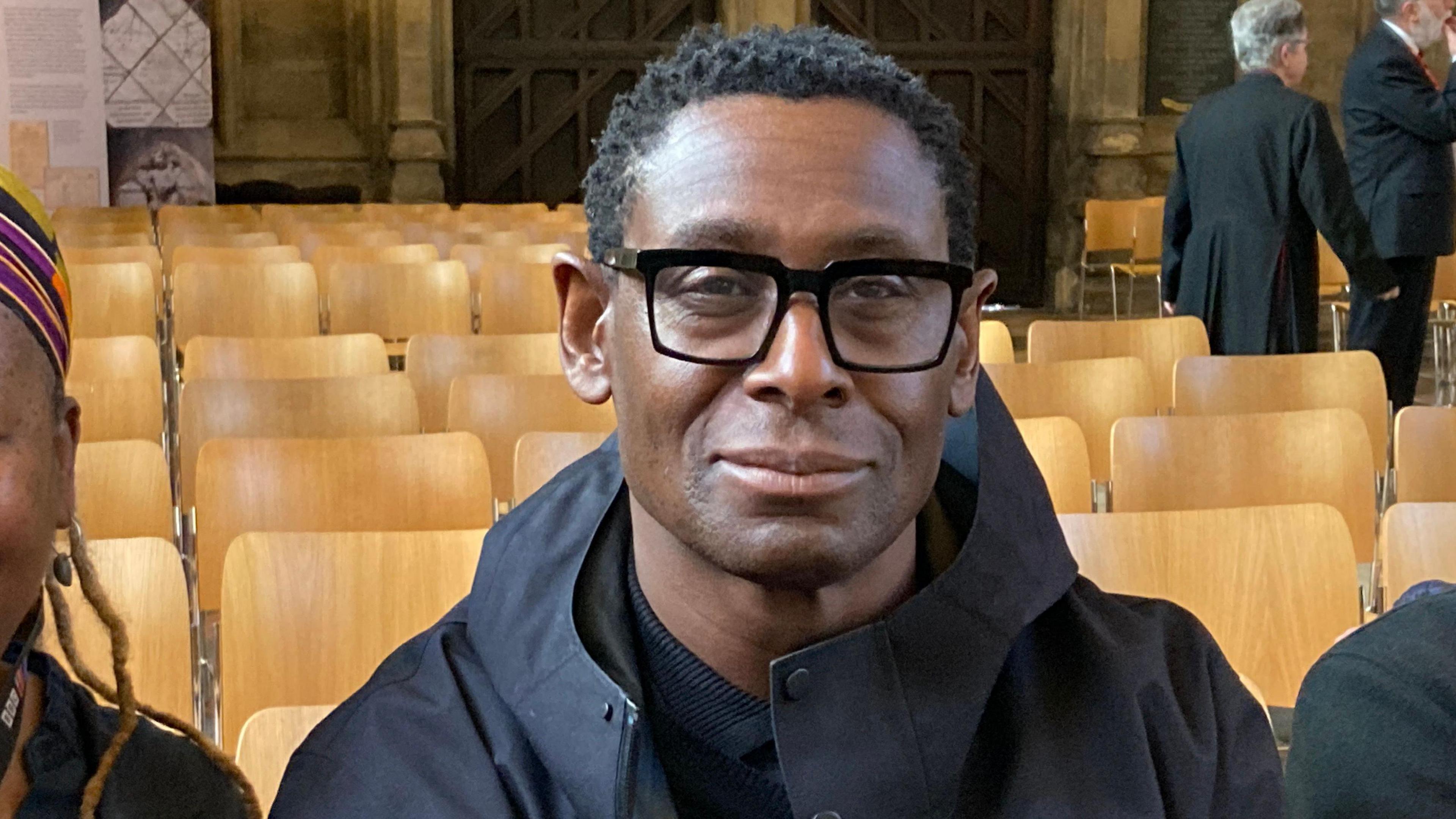 Actor David Harewood is sitting on a chair inside Bristol Cathedral. He is wearing a dark blue coat. He is looking at the camera.