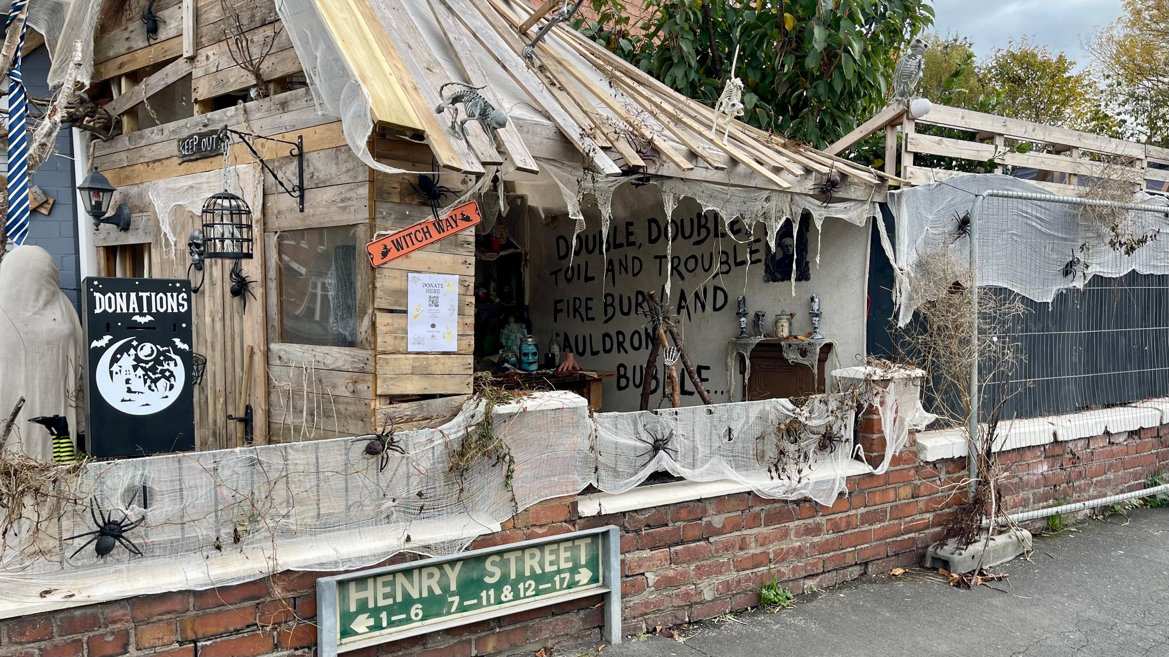 A Halloween-themed wooden shack is covered in cobwebs. It has been built in a garden behind a low brick wall, pavement and green sign reading 'Henry Street.' Inside the shack, the words 'Double, double, Toil and trouble, Fire burn and cauldron bubble...' have been painted on a large sheet.