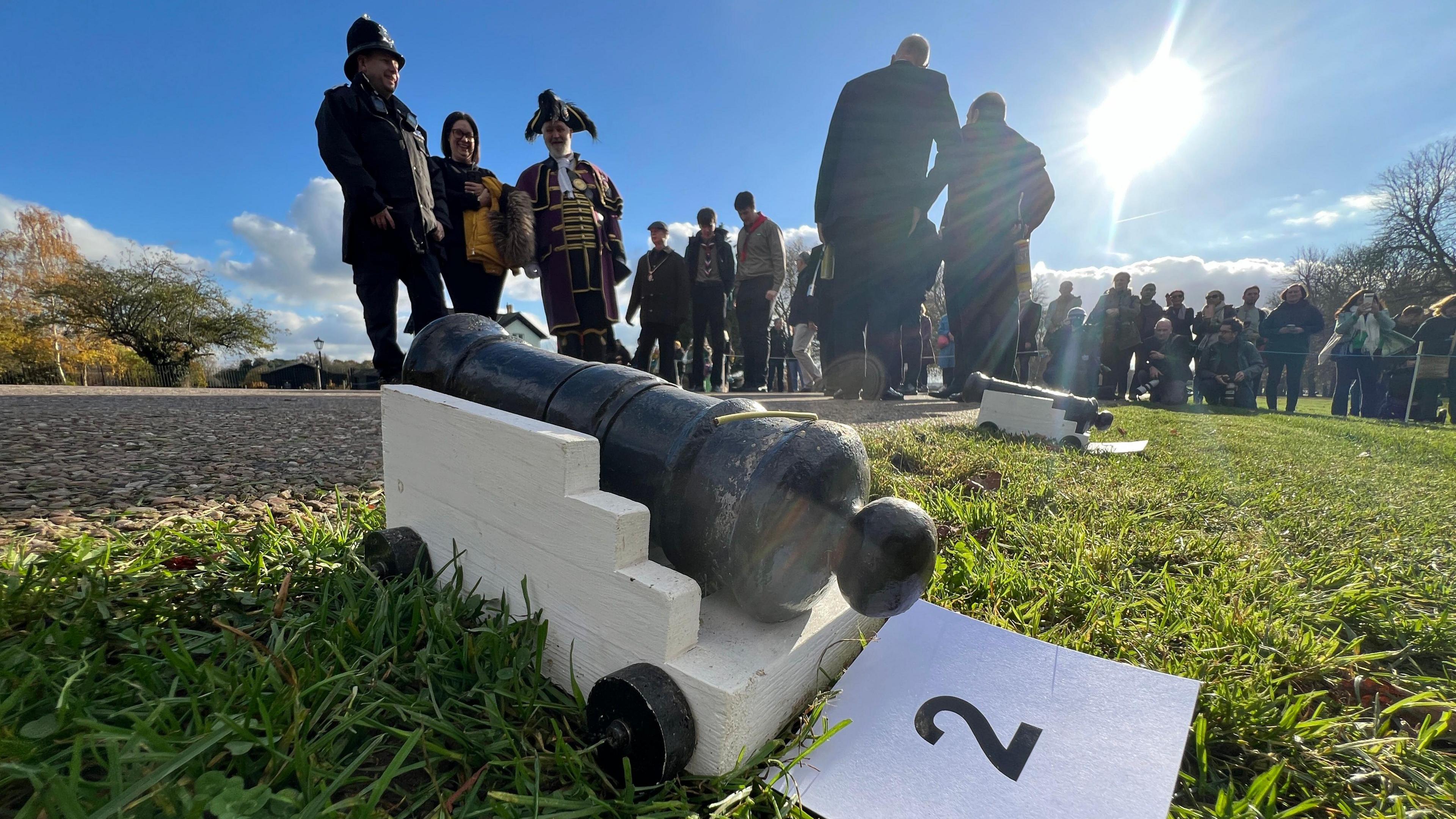 A close-up shot of one of the small cannons on the grass next to the road. There is a crowd of people in the background and it is a sunny day.