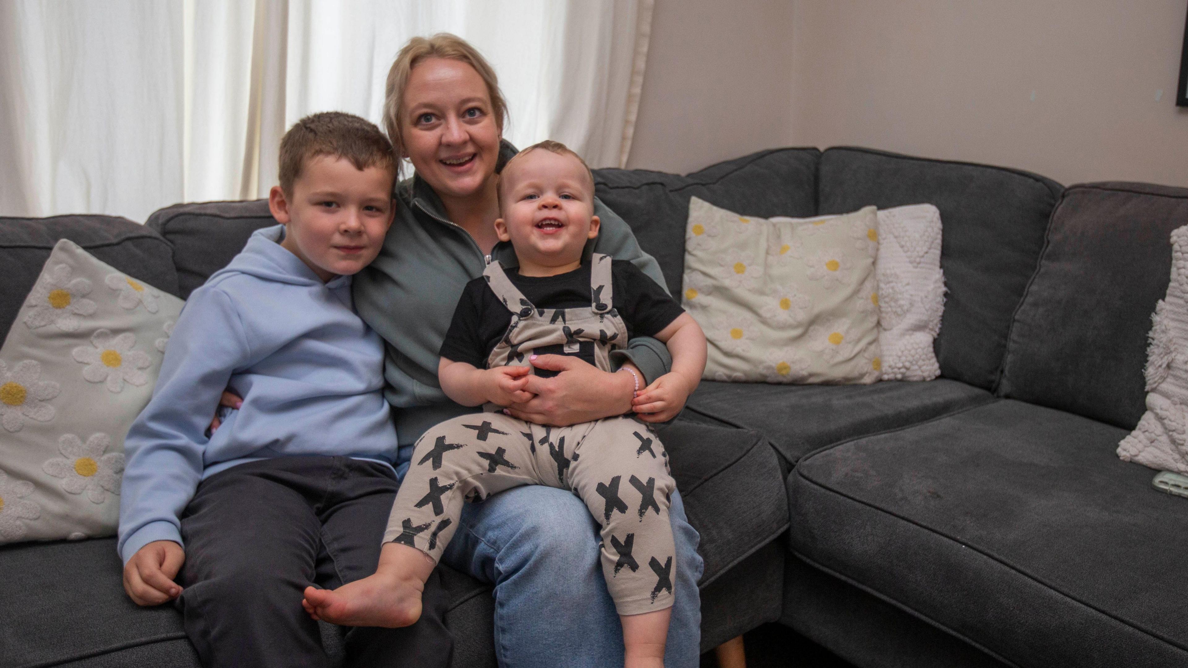 A corner sofar, grey with cushions. On it is a woman with tied-back blonde hair, she has a toddler wearing a black t shirt and black and white dungarees on her lap, and had an around a young boy beside her, with brown haoir, wearing a blue hoodie and grey trousers. They are all smiling at the camera.