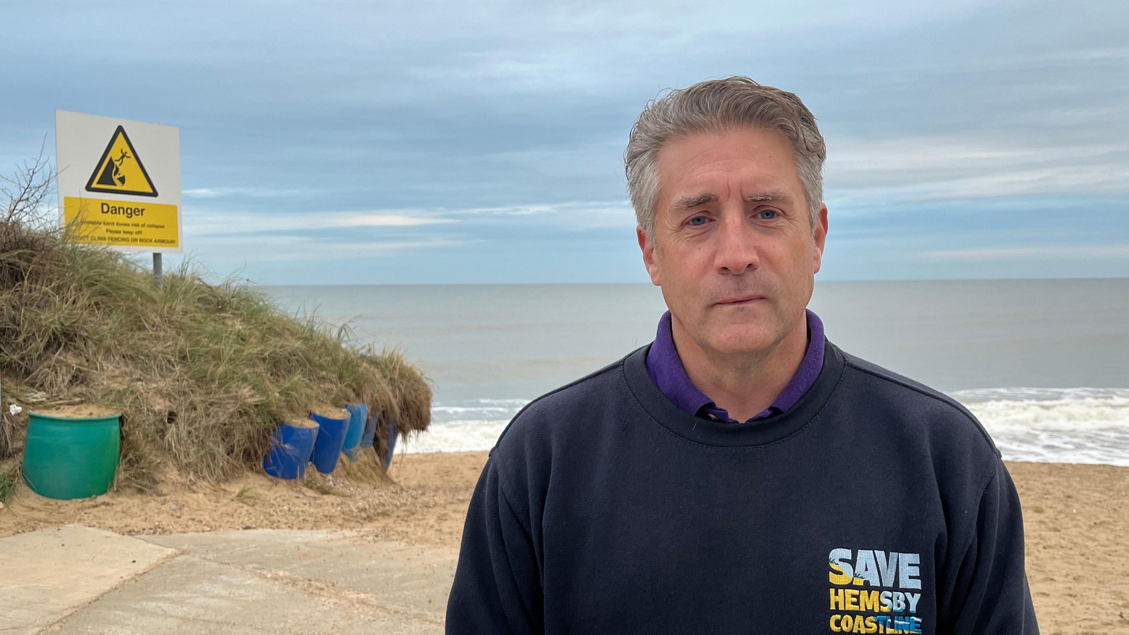 Simon Measures, standing on the sandy beach at Hemsby, with a calm sea behind him. He has sort grey hair and is wearing a blue sweatshirt with save Hemsby coastline written on it. On the left behind him is a dune, covered in scrubby grass and a yellow and white sign saying danger. 