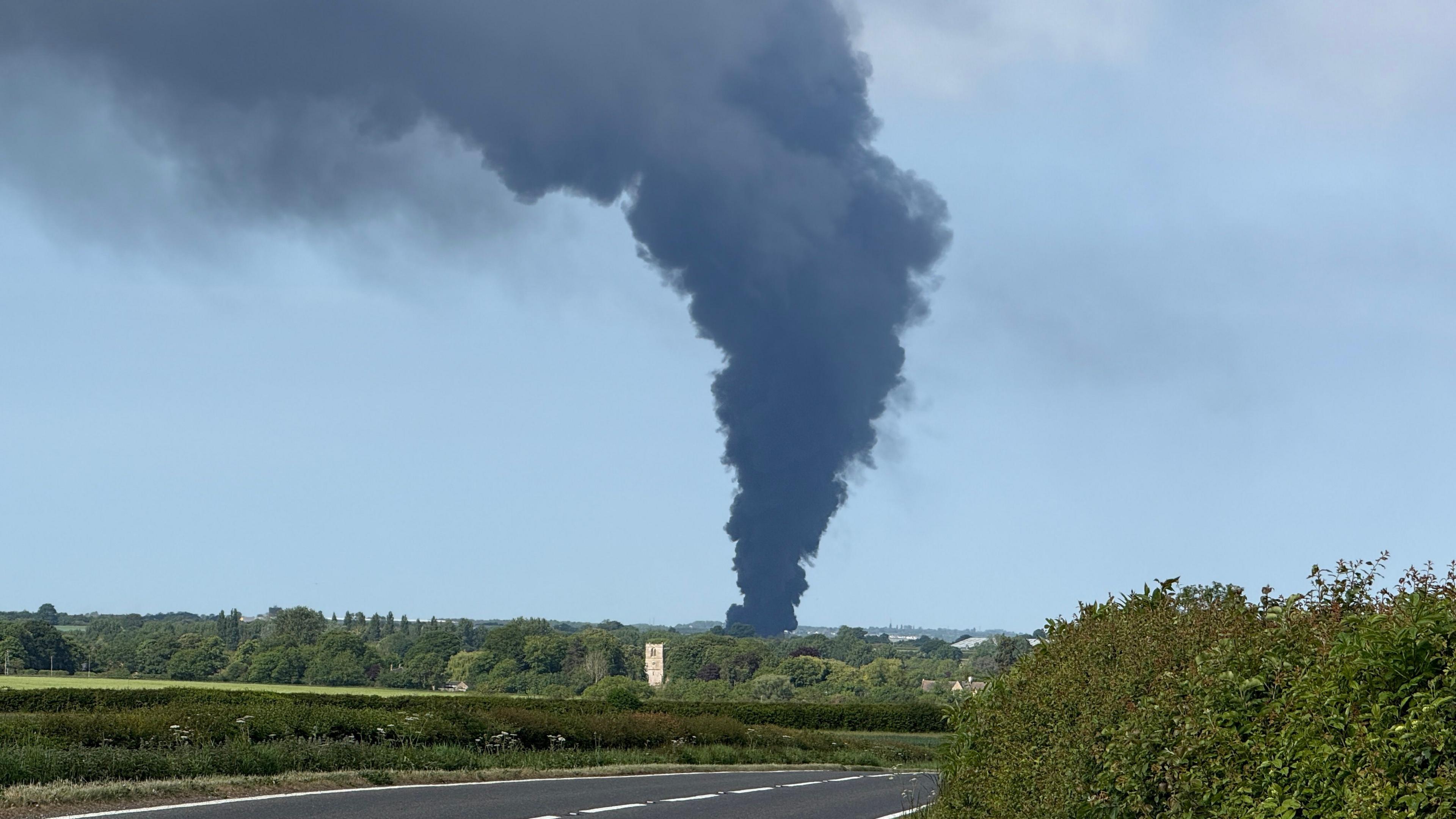 Dark grey smoke funneling out from base to top in the centre of the photo. There are fields with hedges around the smoke plume and a road in the foreground.