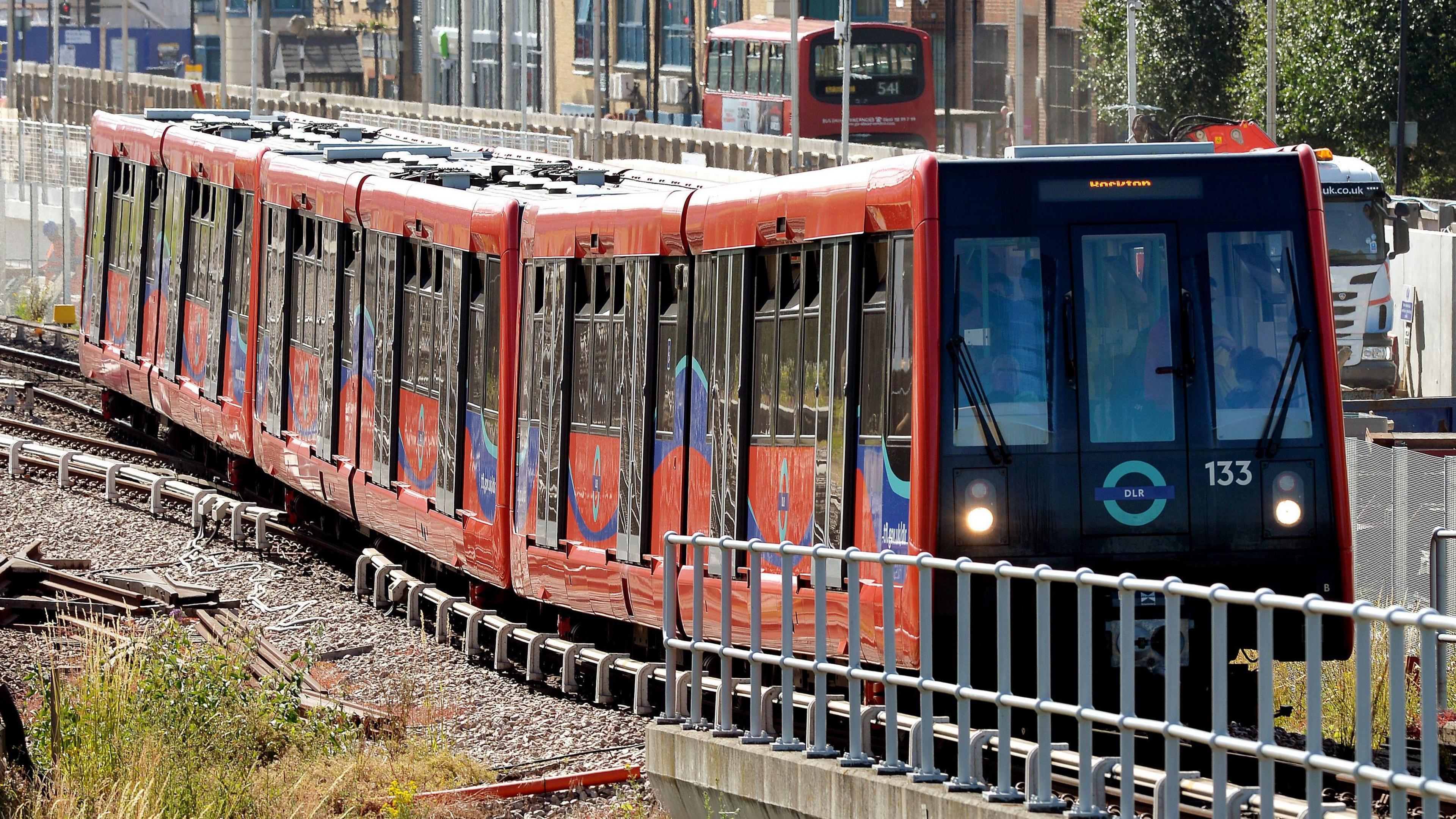 A file image showing a red DLR train travelling on the line  on a sunny day, with a road with a bus and lorry on it in the background