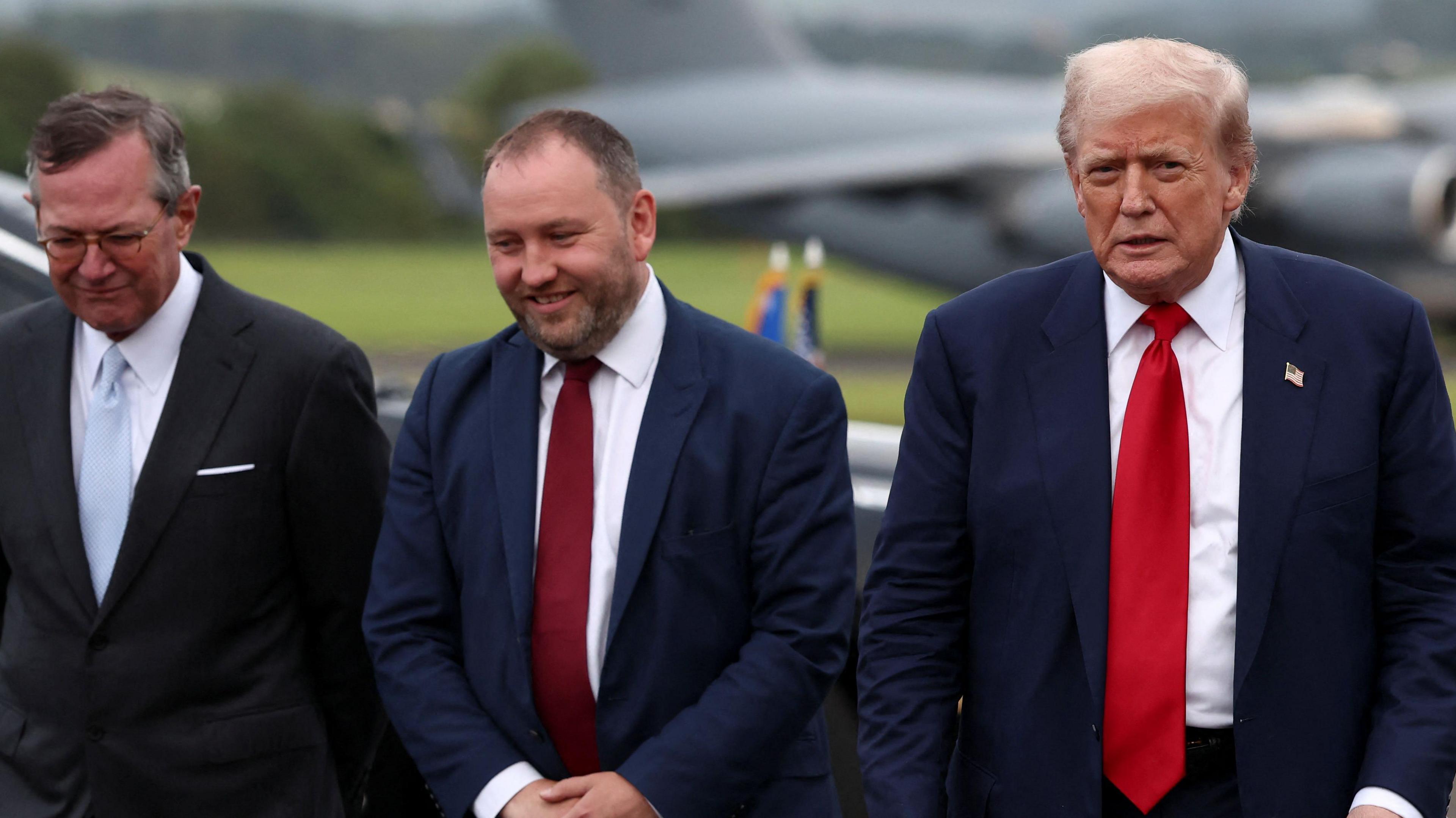 Three men from left to right - Warren Stephens, US Ambassador to the UK, Scottish Secretary Ian Murray and US President Donald Trump. They are at an airport with what is likely Air Force One blurry in the background. All three are wearing suits.