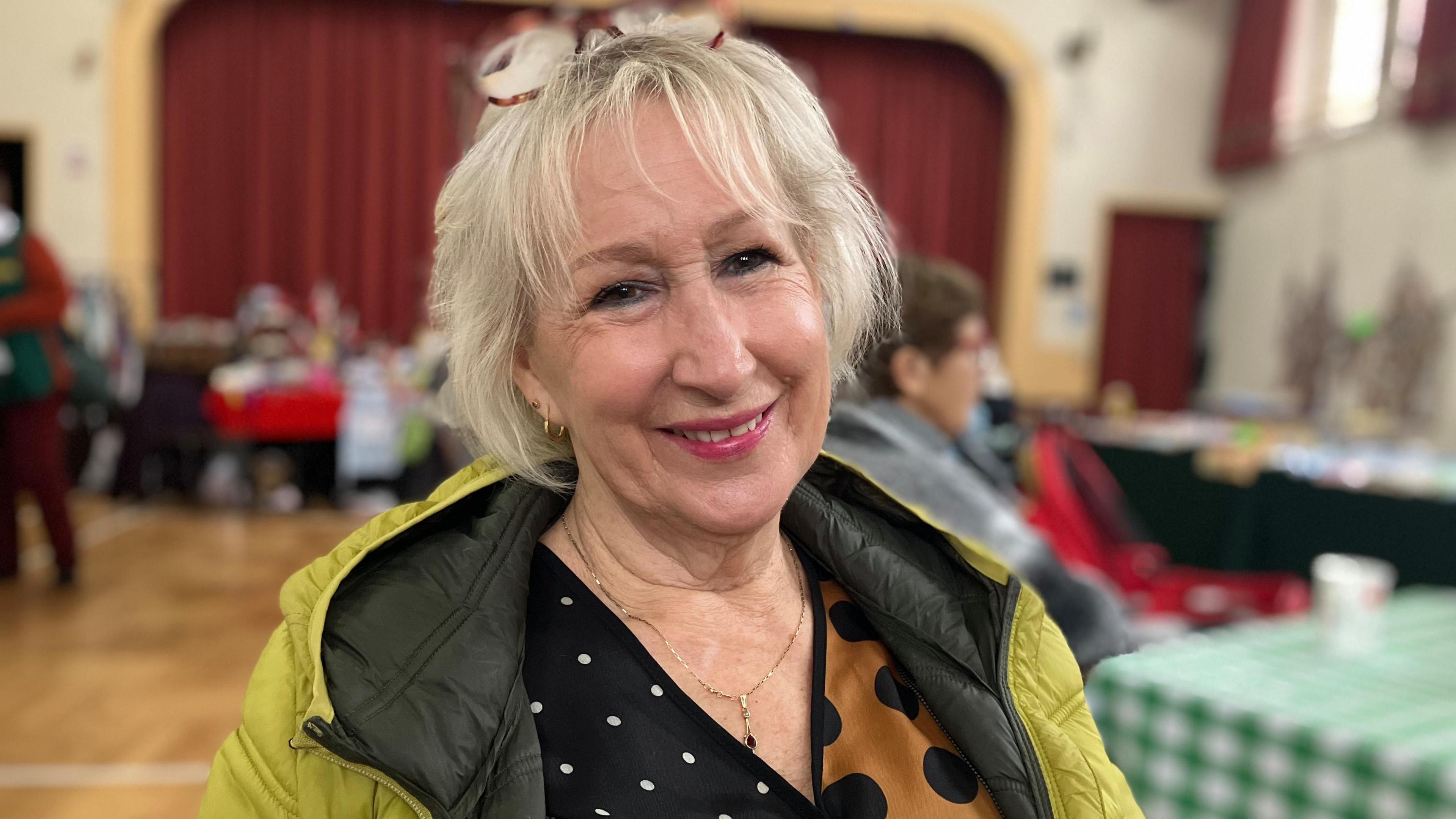 A woman in a village hall smiling at the camera wearing a lime green jacket with glasses pushed back in her hair