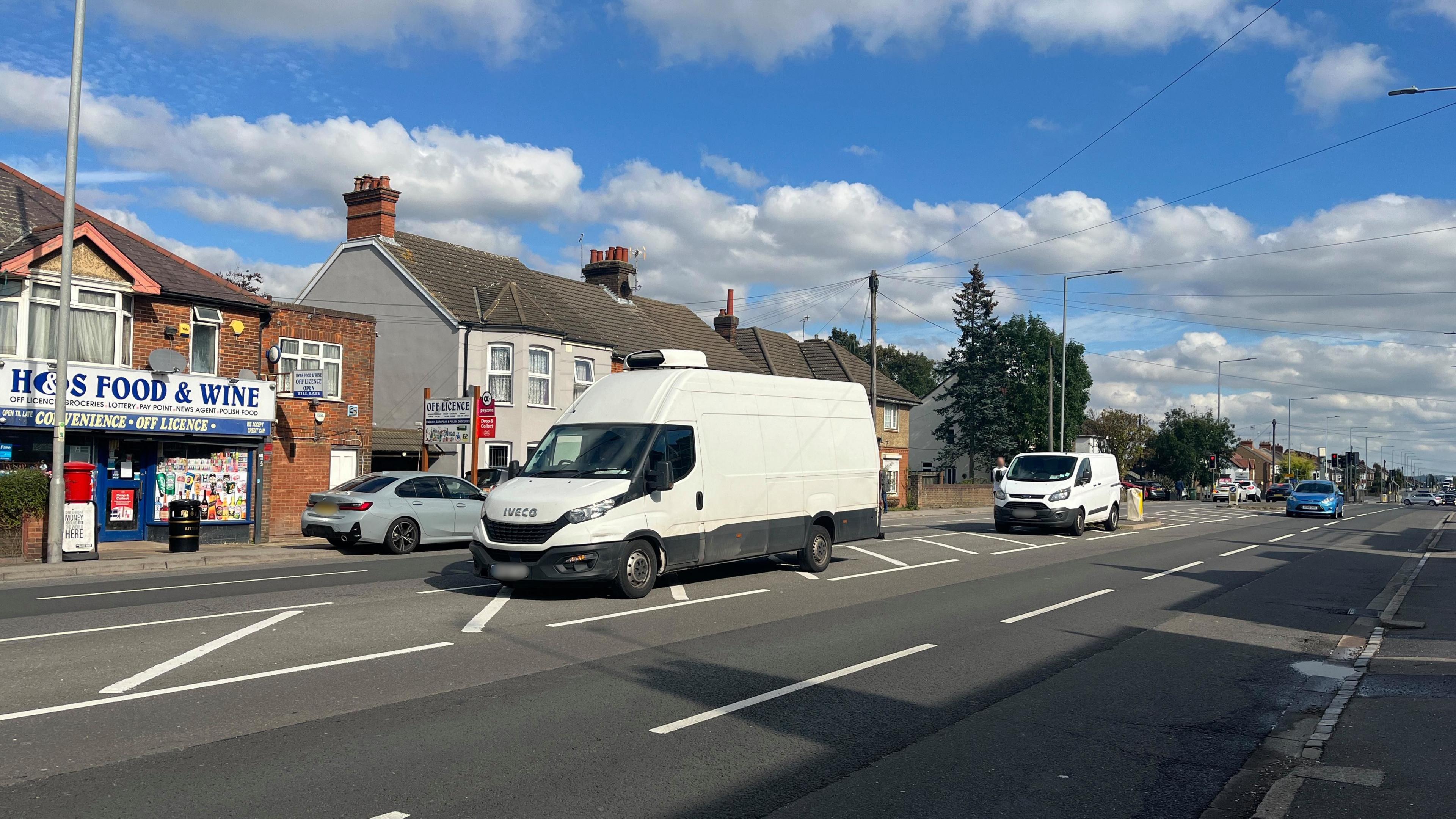 Two vans are parked on the chevrons in the middle of Luton Road in Dunstable with a convenience store on one side among terraced houses.