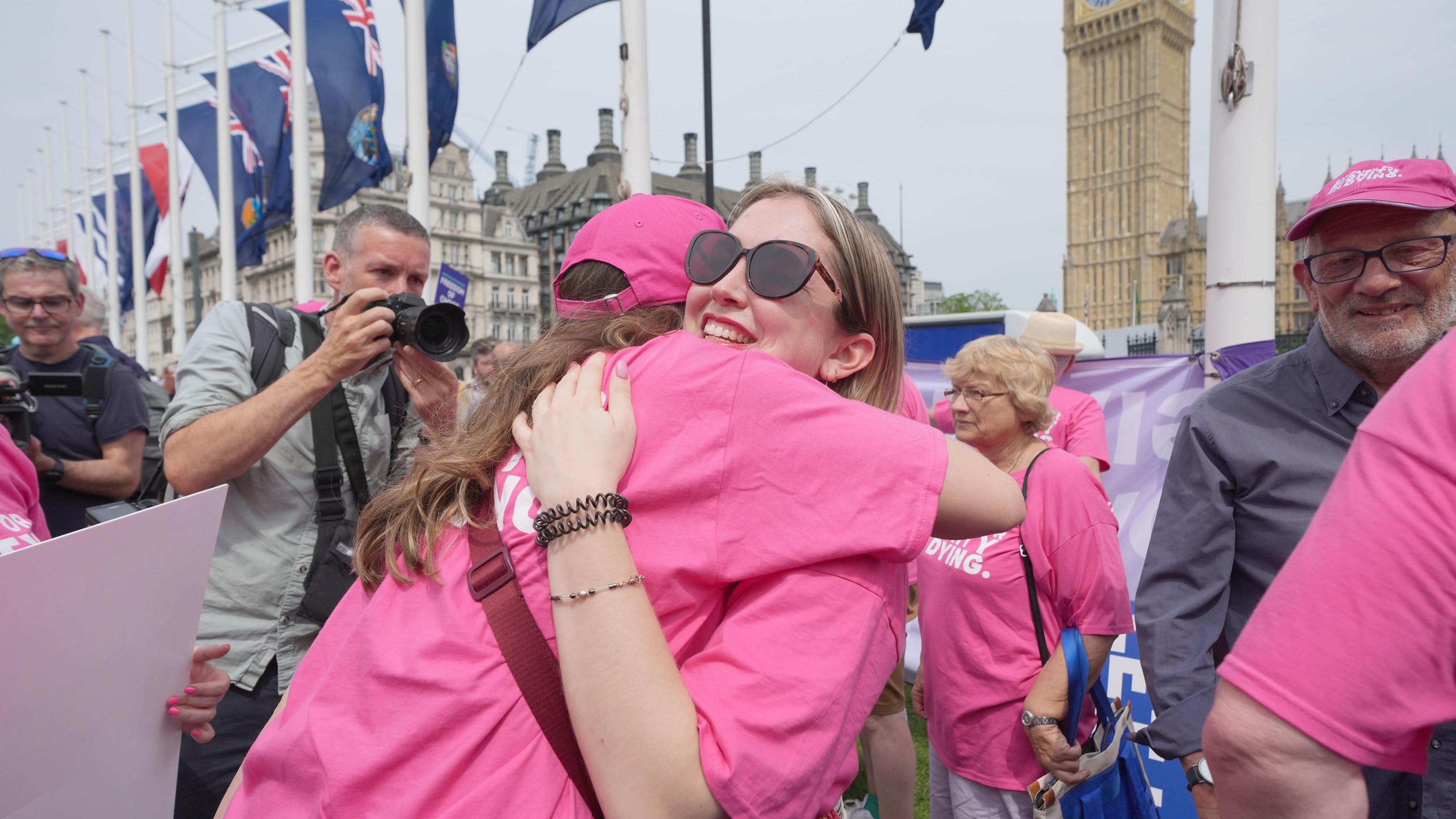 Two women wearing bright pink t-shirts hug in a crowd in front of the Houses of Parliament