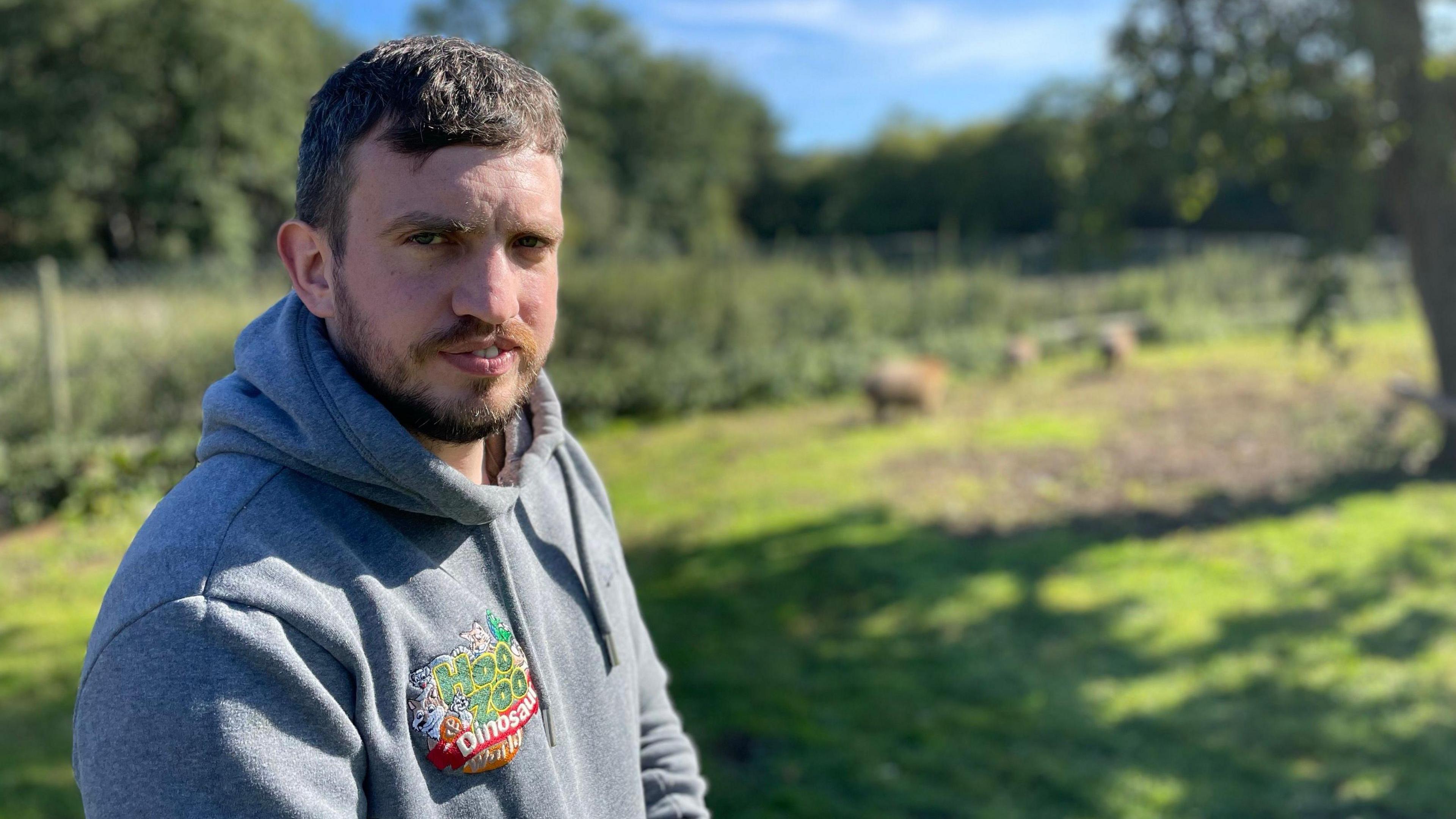 Will Dorrell wearing a grey hoodie with the zoo's logo stands in an area of the zoo with grass and trees behind him. He has short dark hair and a beard