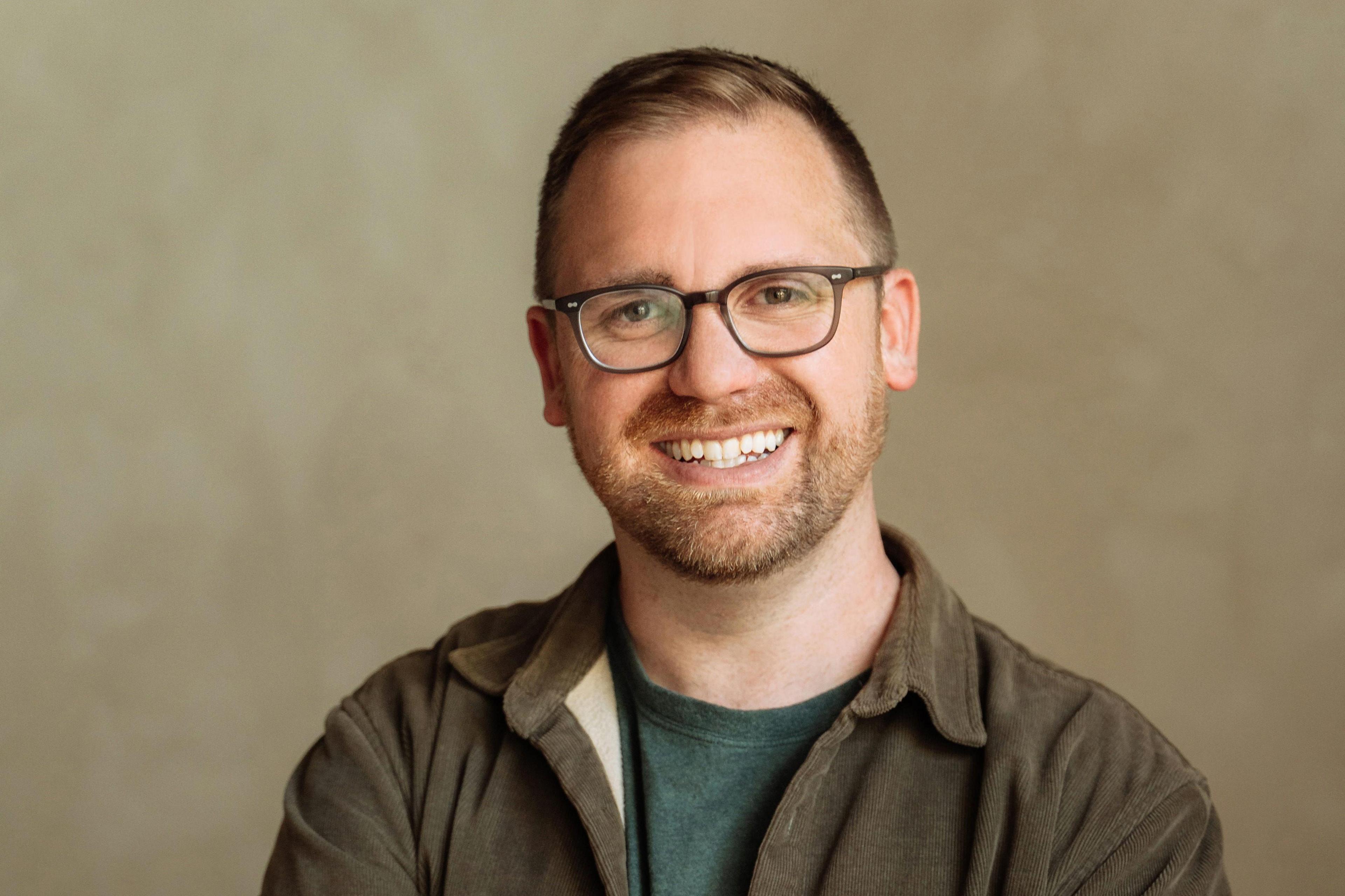 A man smiles at the camera, he is wearing brown glasses and a brown jacket over a green t-shirt. He has brown hair and facial hair. 