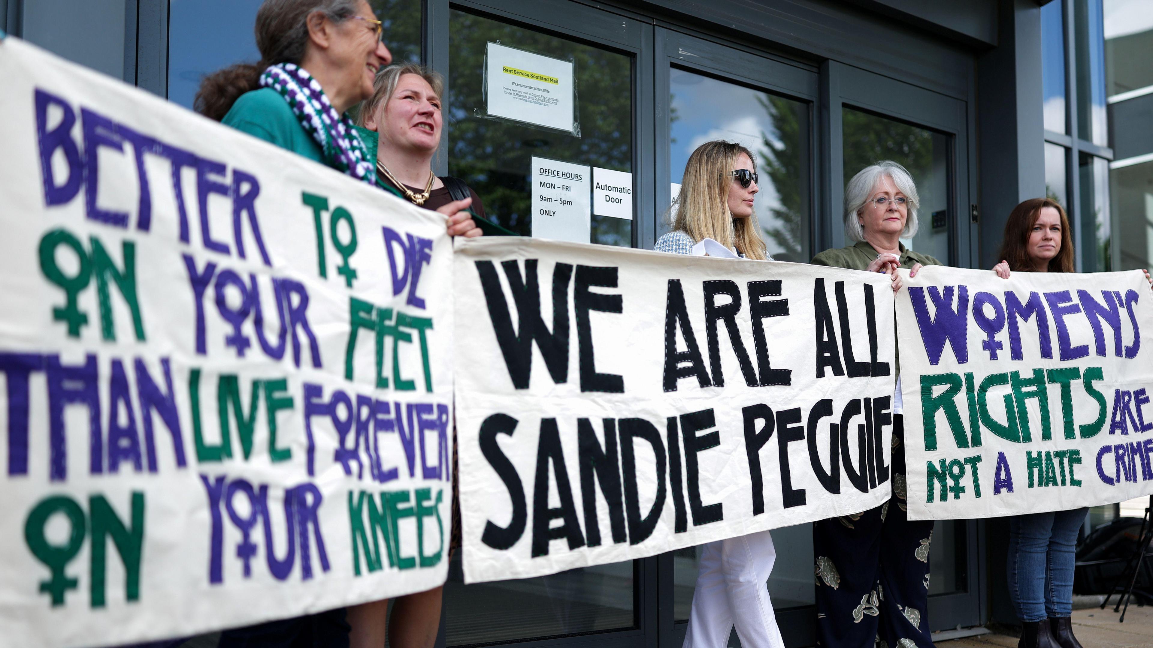 Several women hold banners supporting gender critical nurse Sandie Peggie - the banners saying We Are All Sandie Peggie, better to die on your feet than live forever on your knees and women's rights are not a hate crime