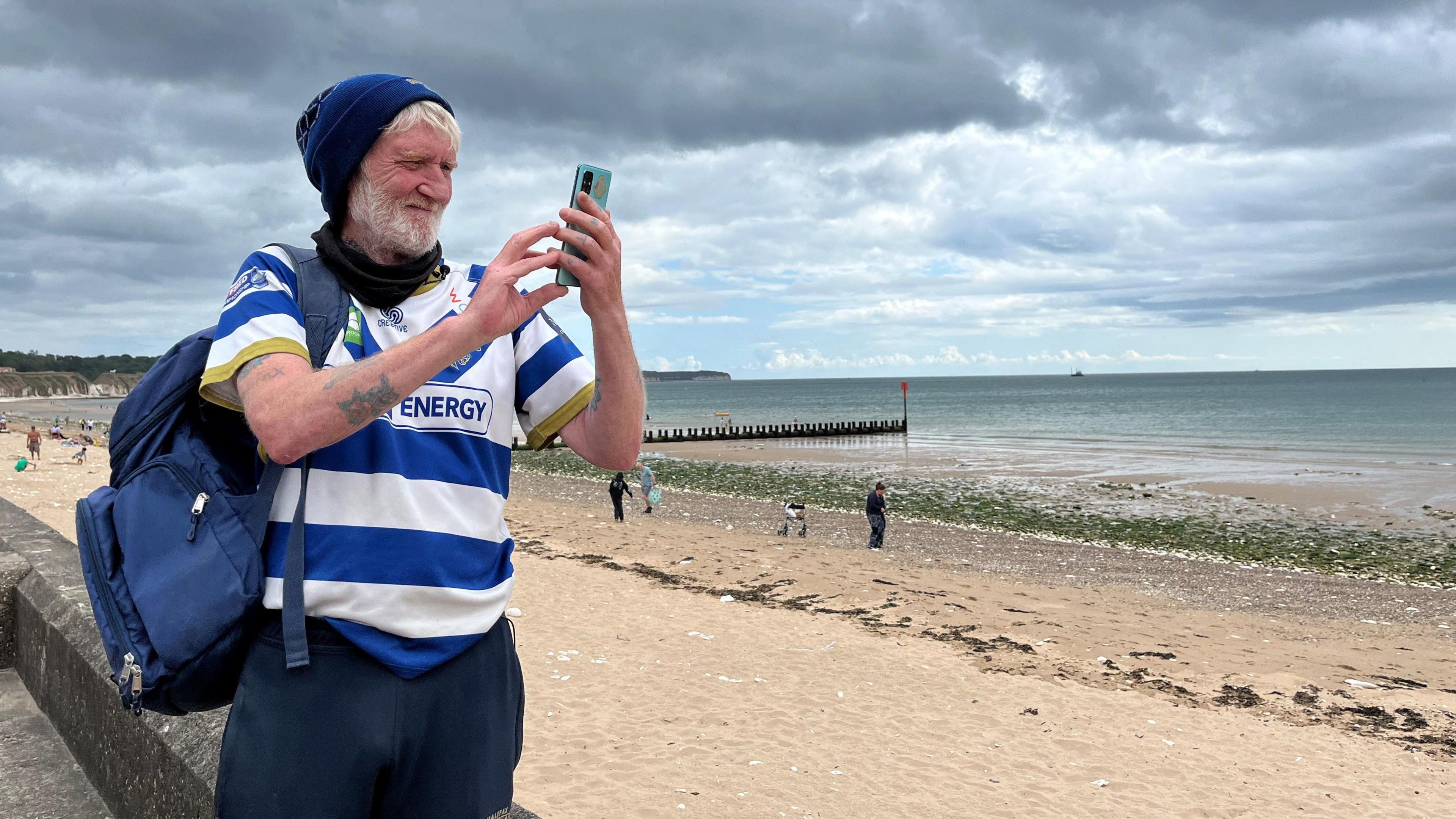 An older man is taking a photo of the beach. He is using an Android phone to do so. He is wearing navy tracksuit bottoms, a blue and white sports shirt (Halifax Panthers) and a navy blue woolly hat. He has a large dark blue rucksack on his back. The majority of the image is of the beach - sand, sea and a sky covered in white and grey clouds. The background is blurred but still visible - the seafront can be seen. 