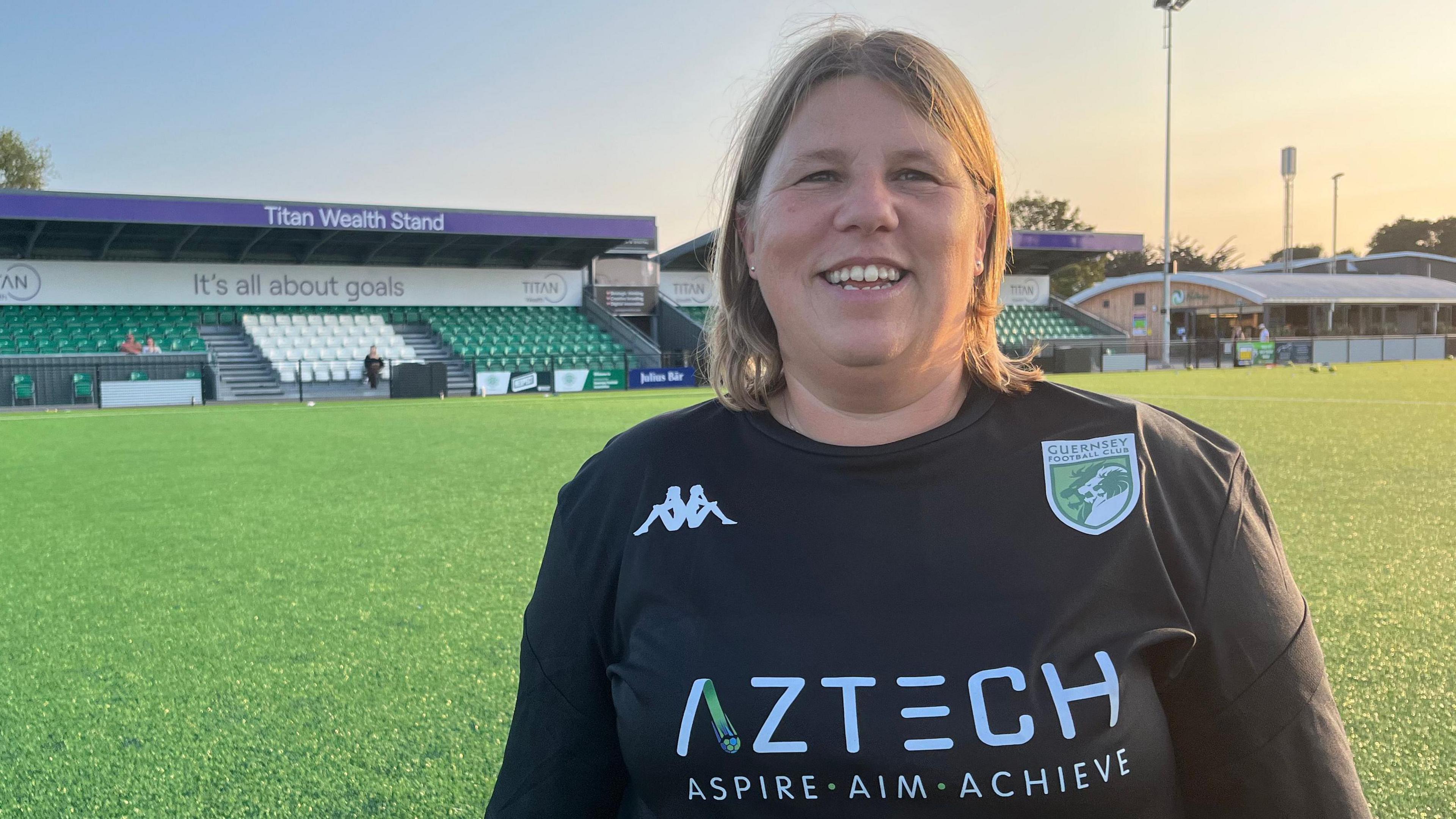 Anna Gauvain smiles at camera with the club's main stand in the background, wearing black training top with club badge