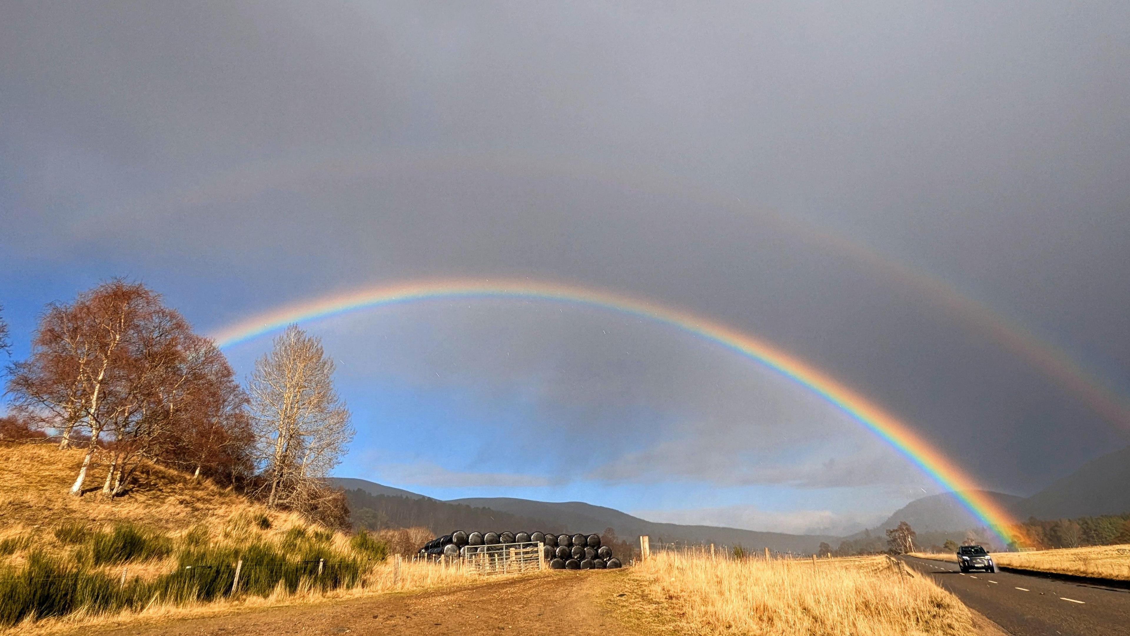 A double rainbow over a road in Aberdeenshire. The outer rainbow is against a grey sky and is very faint. The inner rainbow is much brighter. The sky above it is grey but below it is light blue. The grass below is golden and straw like. A tarmac road can be seen to the right side while there are trees on the left side.