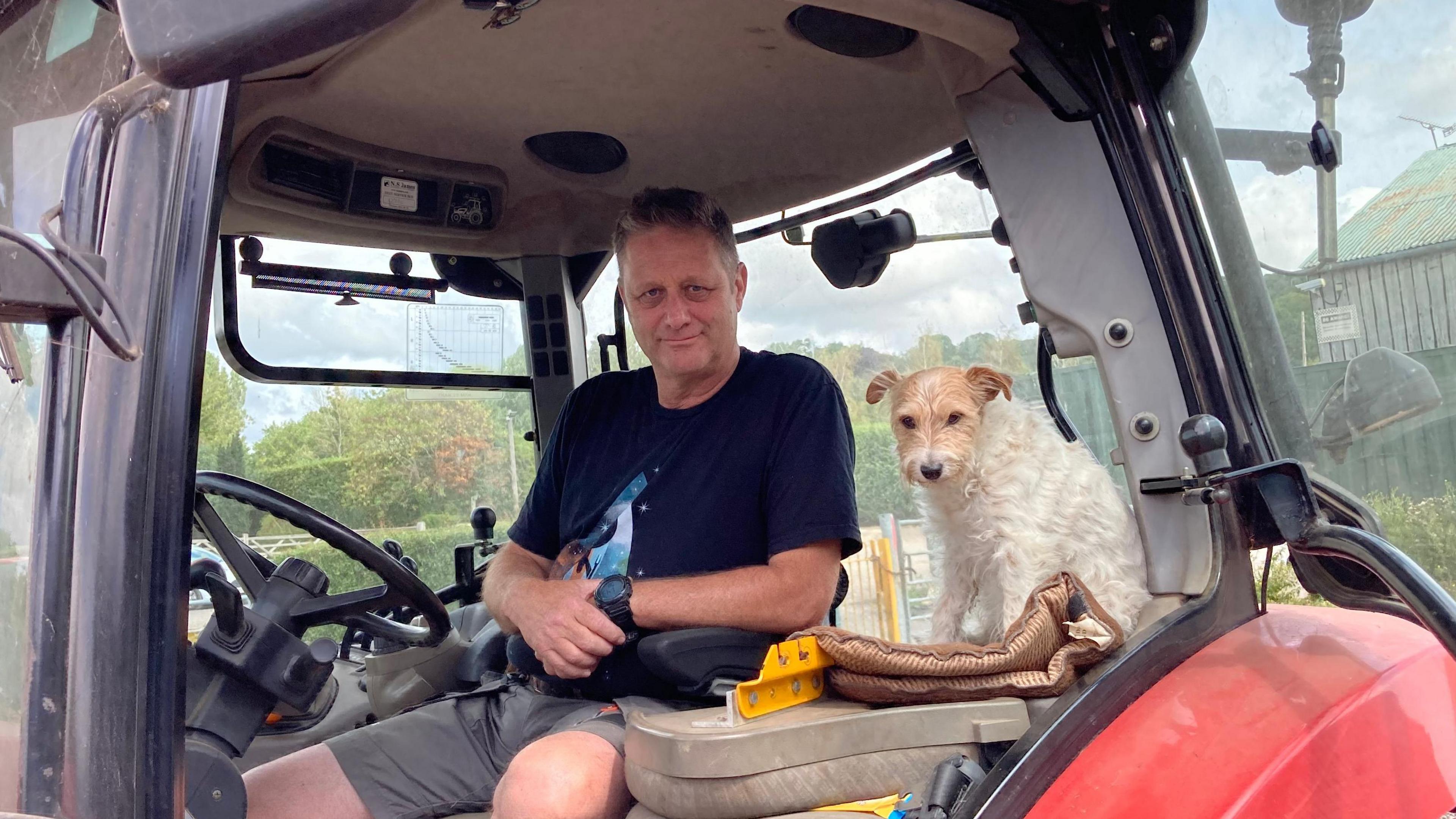 Ian Pullen sits at the wheel of a red tractor with dog Biskit, a parson's terrier, sitting next to him. Ian is wearing a black T-shirt and shorts, Biskit has white fur with light brown fur on his face.