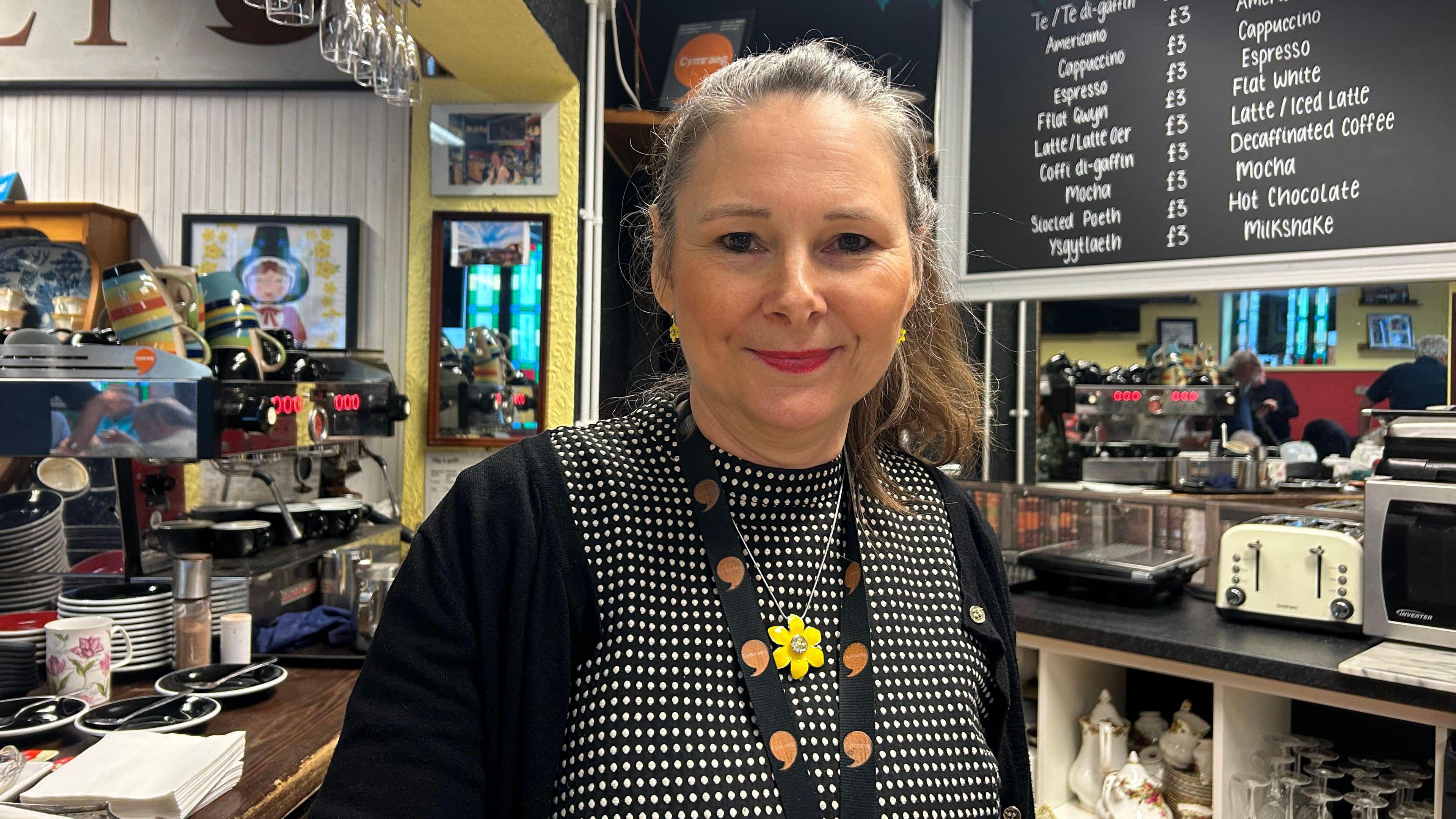 Teleri Jones mewn crys du a Gwyn a chardigan ddu. Yn gwisgo Lanyard siarad Cymraeg a mwclus gyda blodyn melyn.