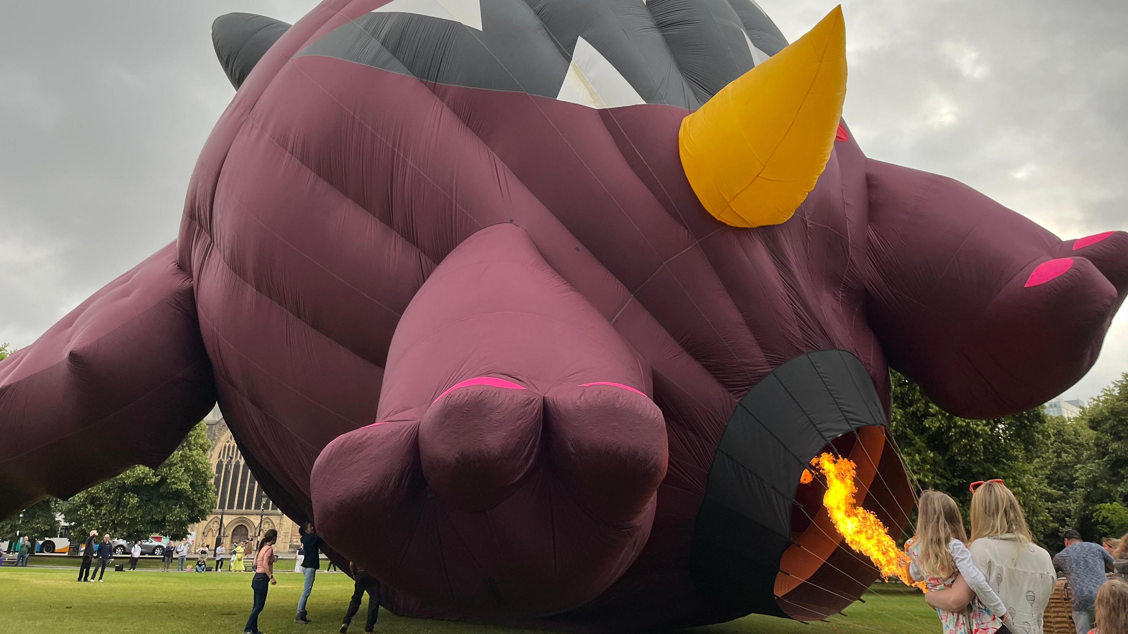 A purple monster shaped balloon being inflated using fire. A mother and child watch.