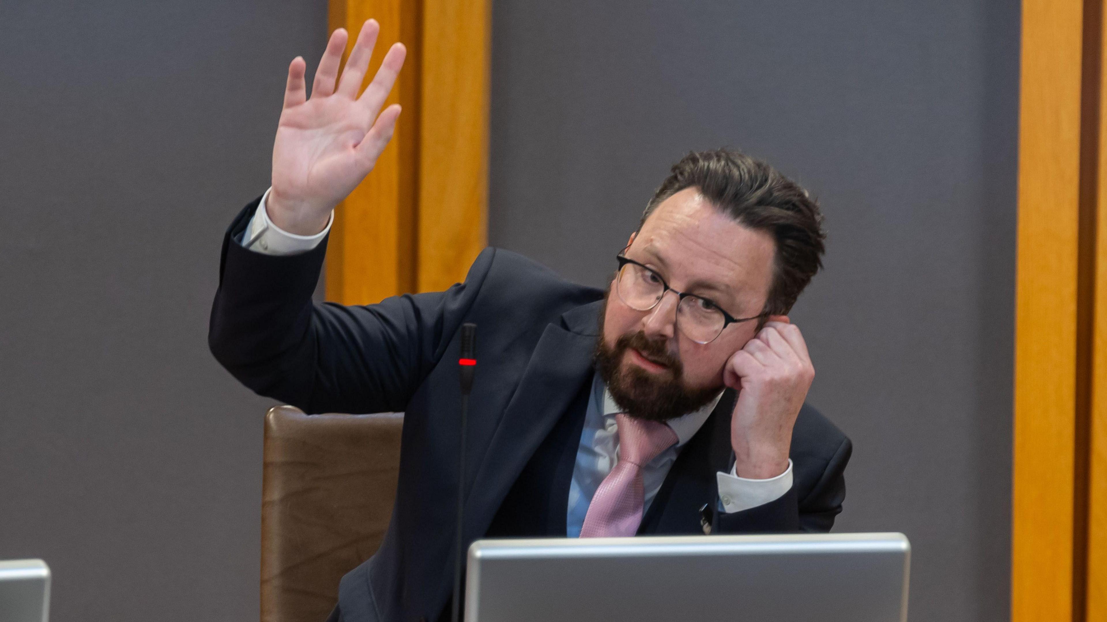 Hefin David sat in the Senedd debating chamber with his left hand to his ear and his right hand up, gesturing to be called to speak. He is wearing a dark suit, blue shirt and pink tie, has a beard, dark hair and glasses.