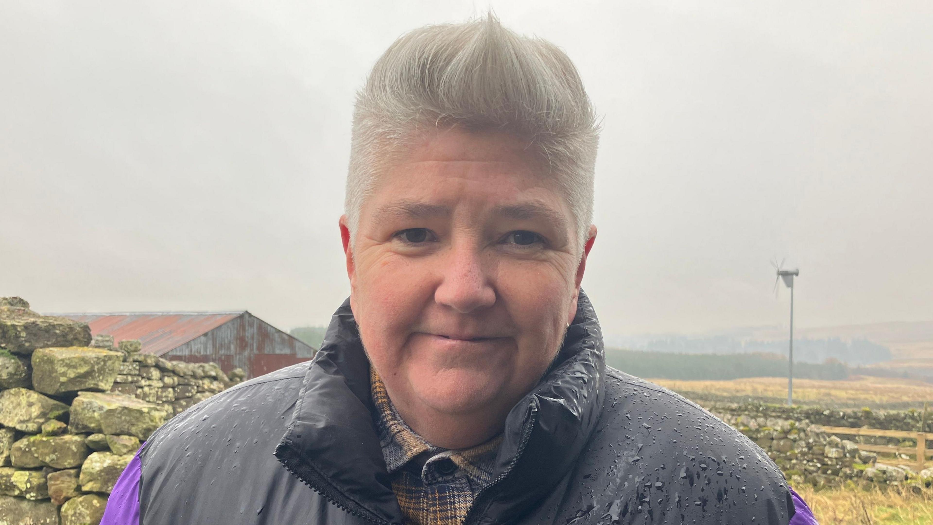 Huffty McHugh is standing in front of a dry stone wall on a grey day on a farm in Northumberland.  She has short blonde hair. There is rain on her the black shoulders of her jacket, the sleeves of the jacket are purple. A farm building and a small wind turbine can be seen behind her in the mist. 