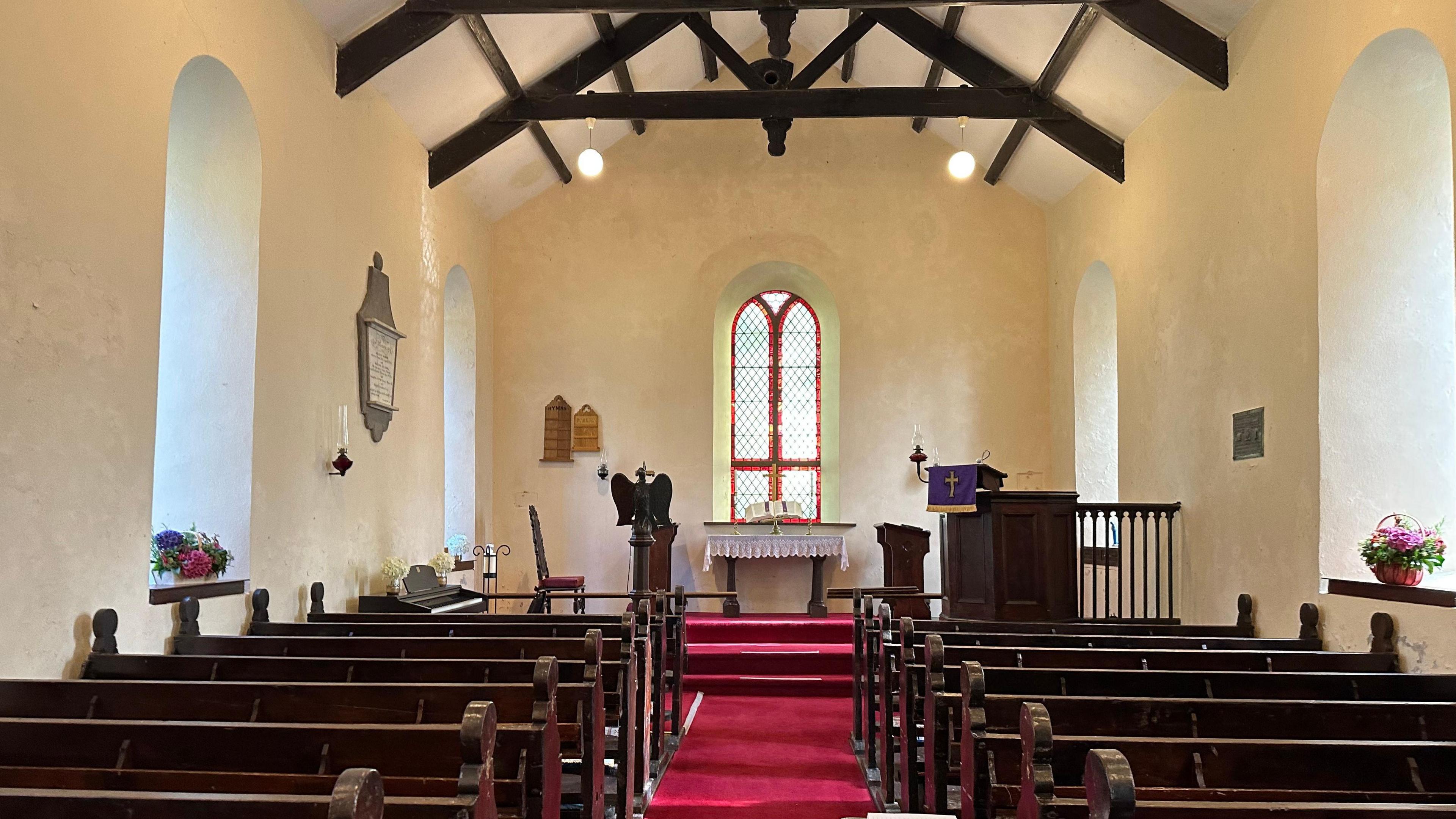 The interior of the church, which is a small, simple church hall, with dark wooden pews, red carpet, arched windows and wooden beams.