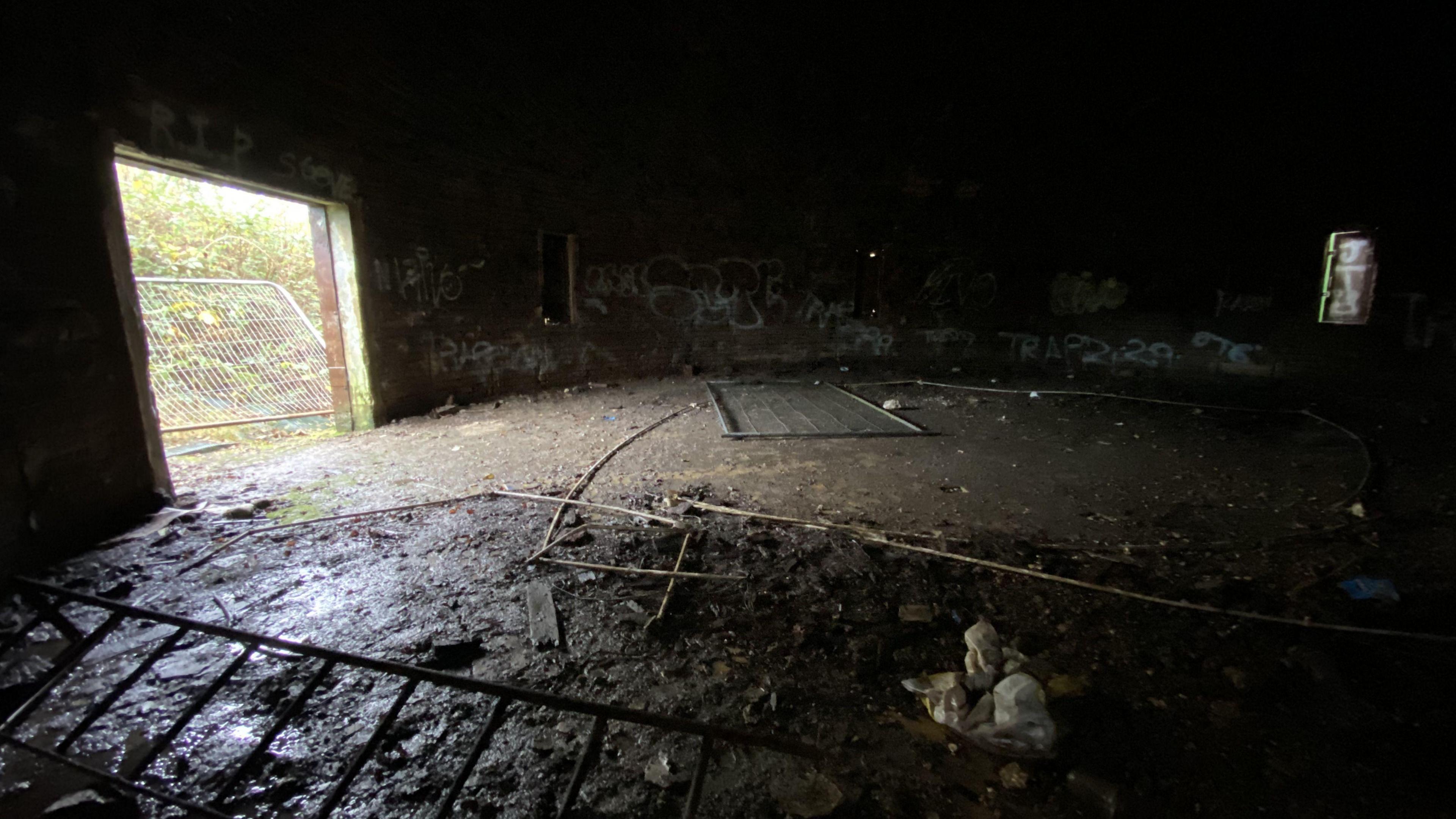 A dark room with mud and various pieces of metal on the floor. There is a large square hole in the left side of the building, which is letting in light