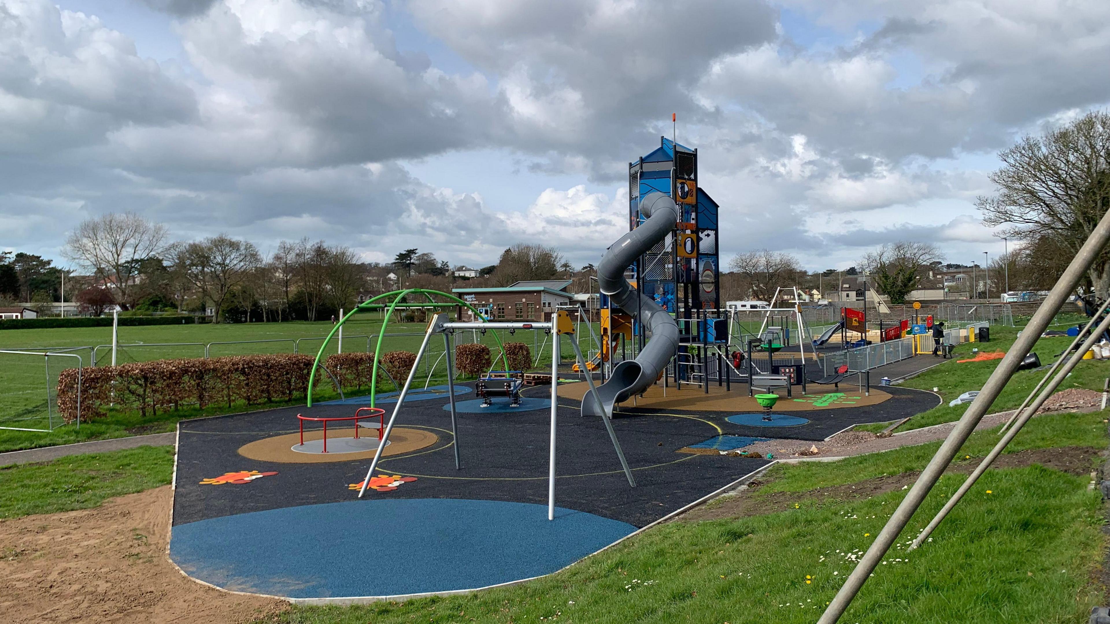 Play park with a tall structure with a slide. There is other equipment including swings and climbing frame in the centre of the park on Tarmac next to a big field.