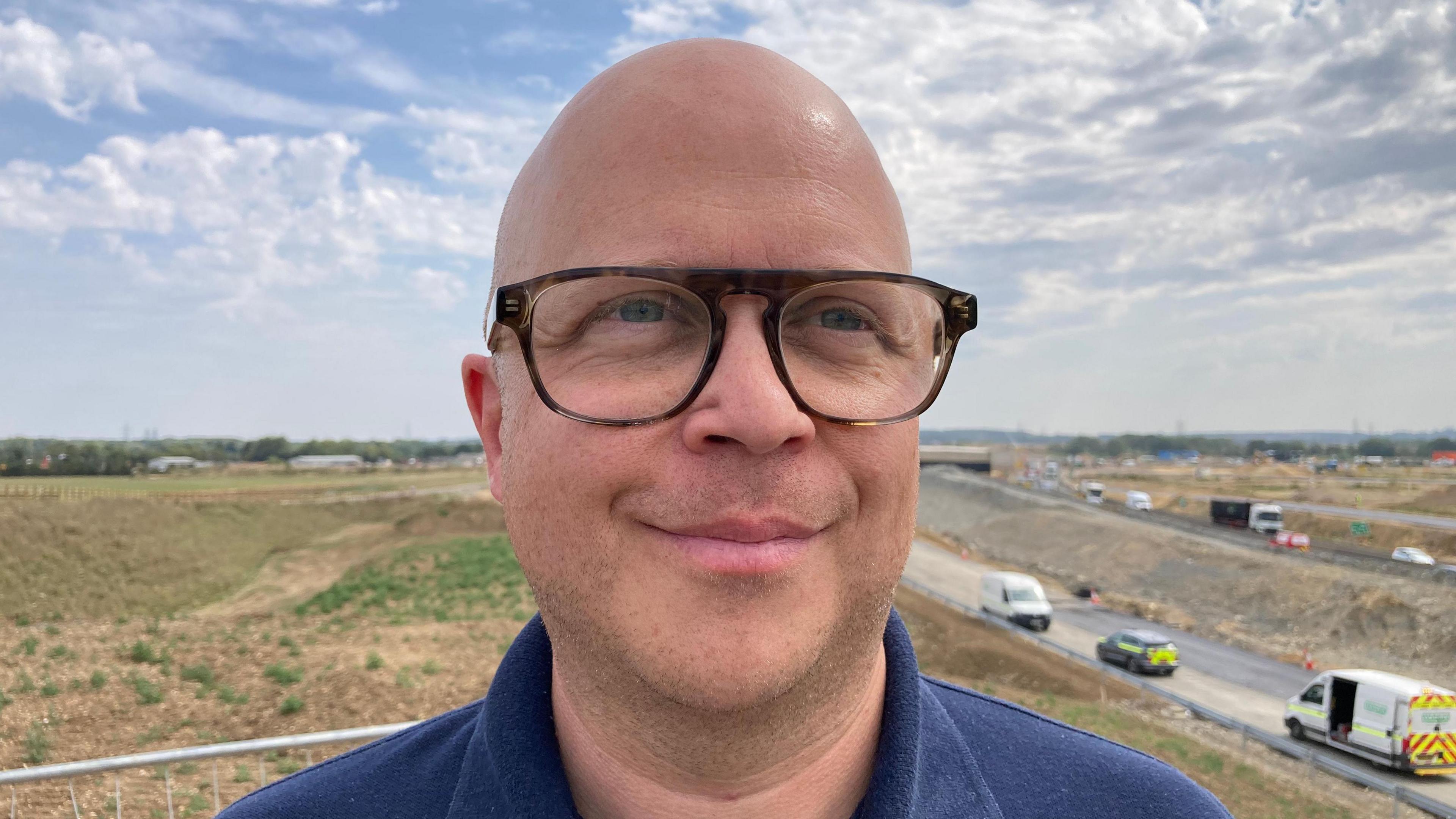 A bald man wearing glasses and a blue top looking to one side of the camera. He is standing on a gantry overlooking roadworks which are in the background.