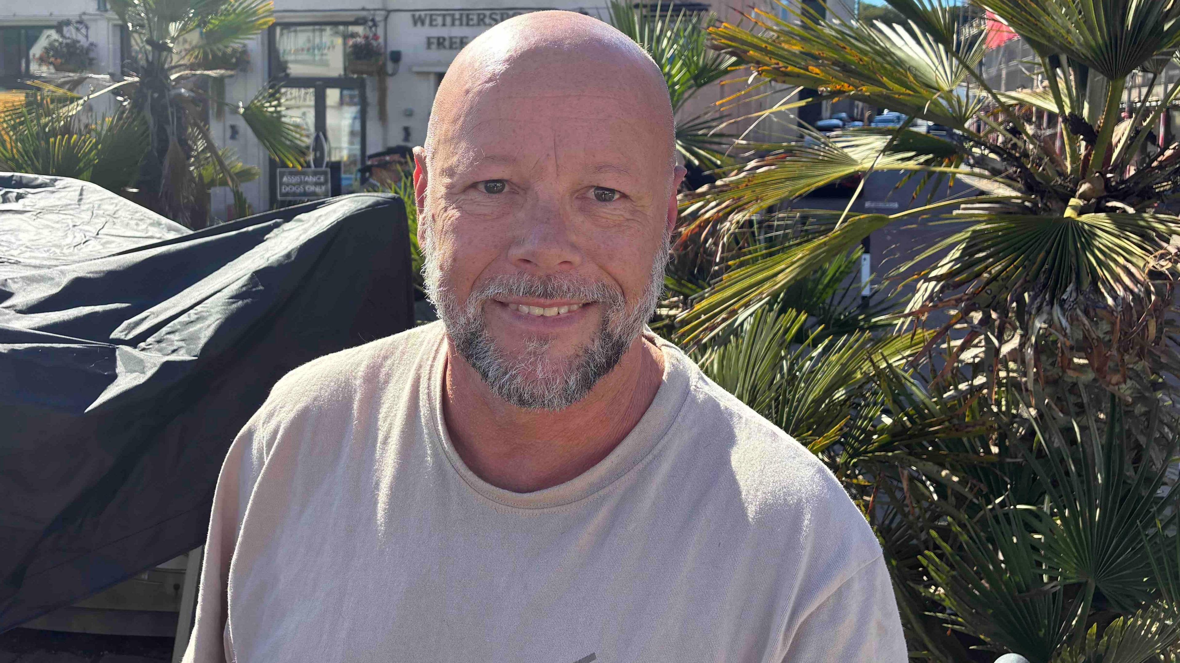 A man with a pepper and salt beard and wearing a light coloured T-shirt is smiling. He is standing besides a palm tree.