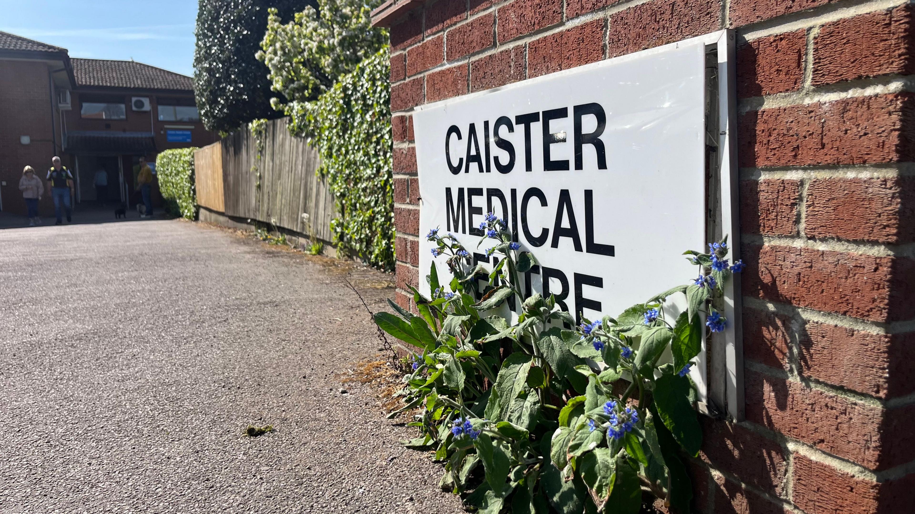 A photo of the Caister Medical Sign, partly obscured by blue flowering borage. It is a sunny day and the practice is visible in the background.