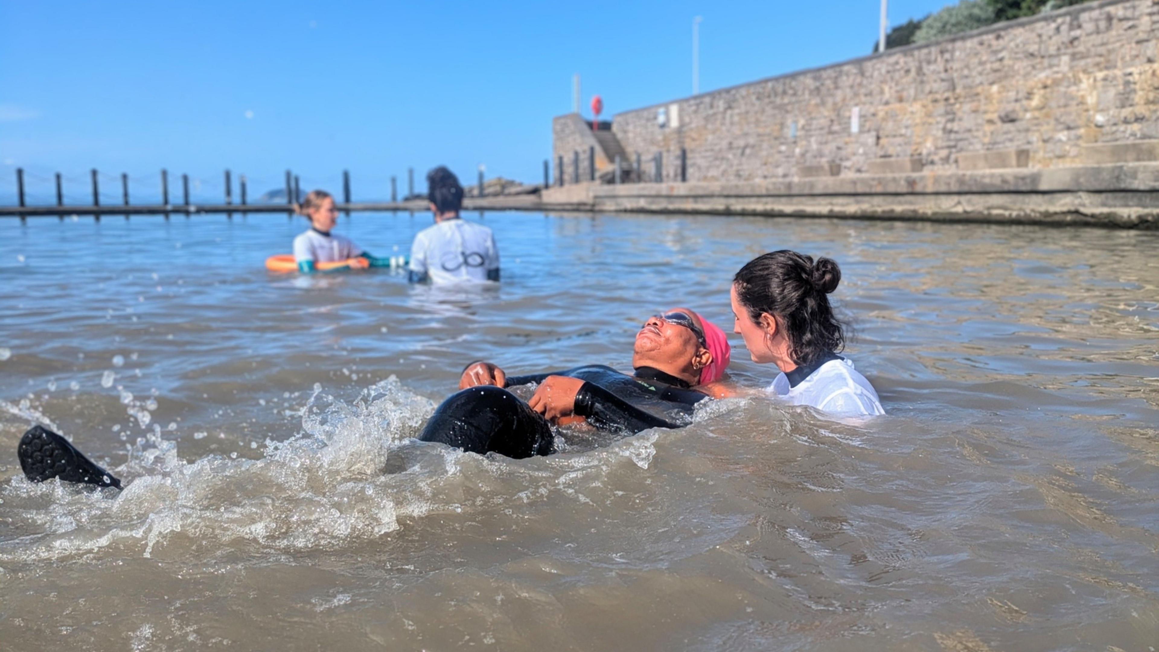 A woman wearing a black wetsuit, pink swimming cap and goggles. She is lying on her back in Weston-super-Mare Marine Lake while another woman holds the back of her head to help her float 