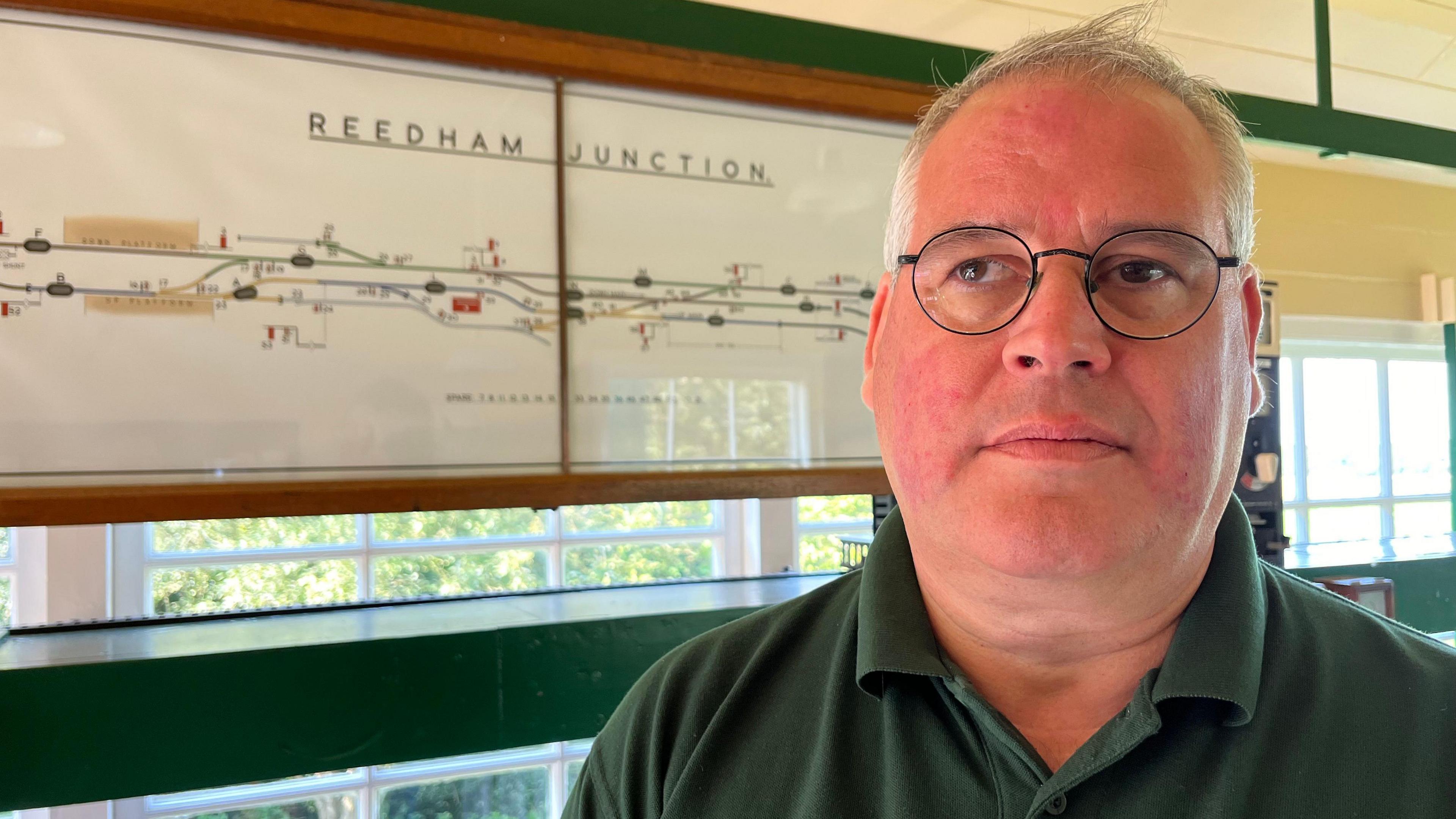 Steven Ashling standed in front of a Reedham junction railway line sign and is wearing circular glasses and a green polo shirt.