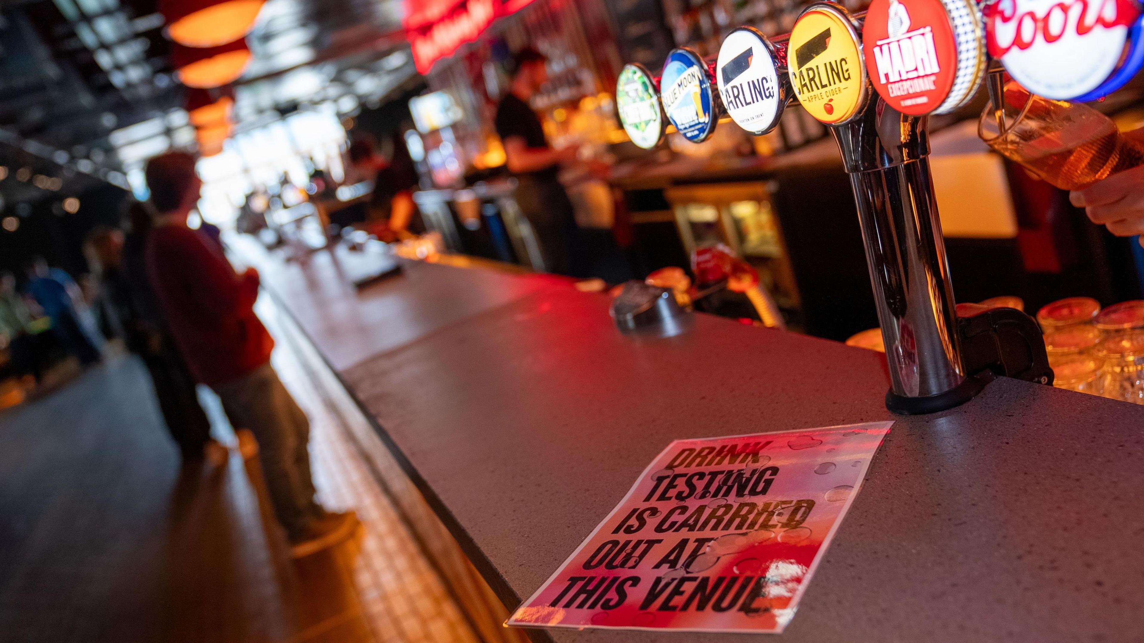 An image of a bar with numerous beer pumps and a "drink testing is carried out at this venue" leaflet.