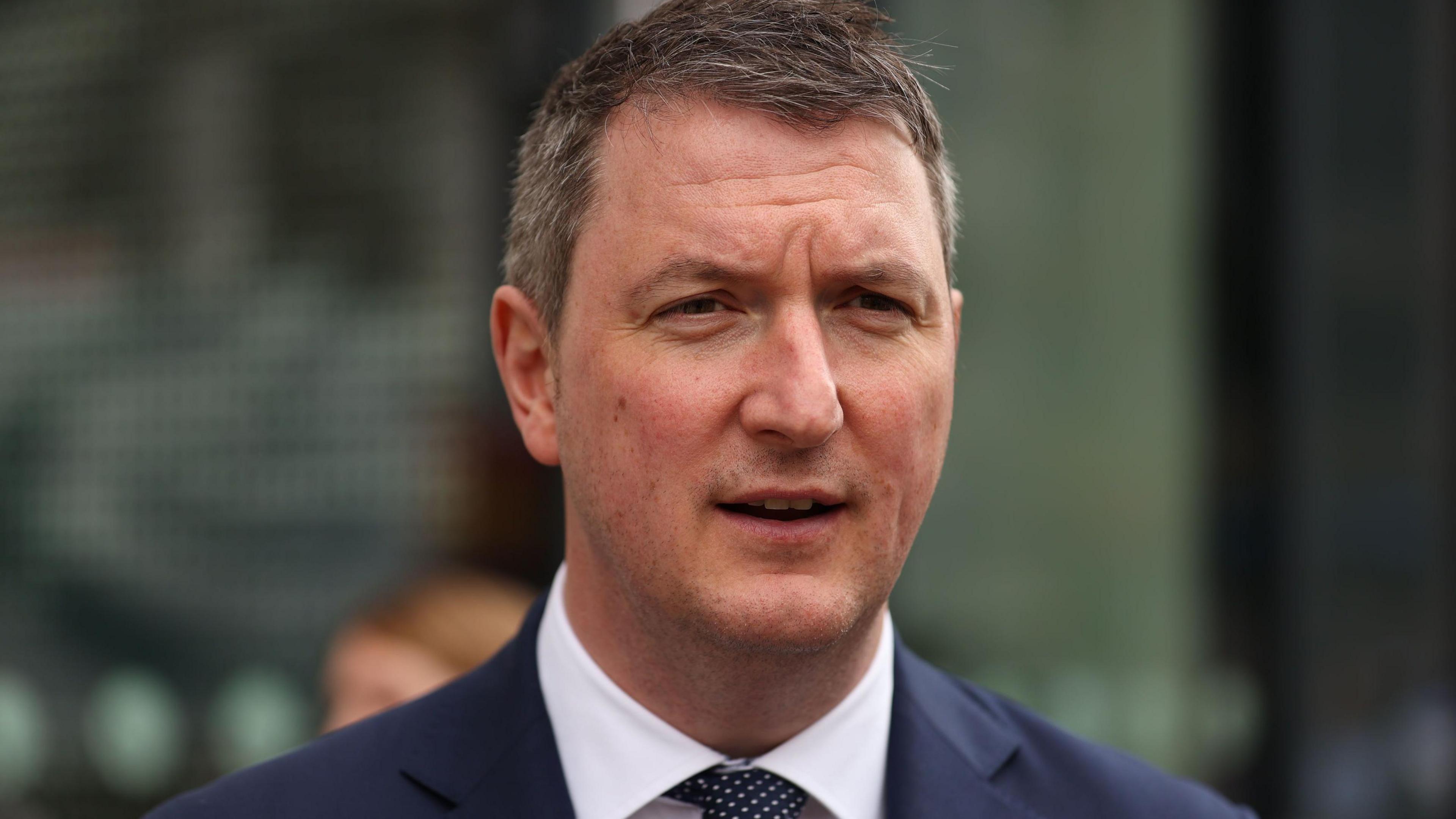 A close up of John Finucane in a dark blue suit, white shirt and black and white tie