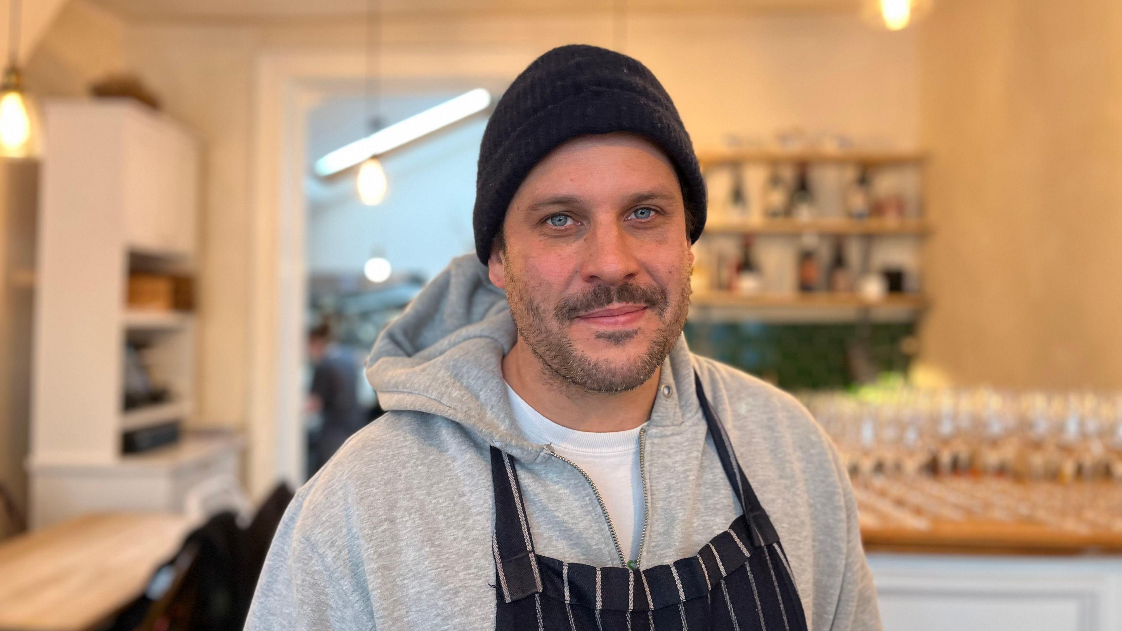 Jan Ostle, wearing a grey hoodie, blue and white chef's apron and navy hoody, standing in a restaurant, smiling