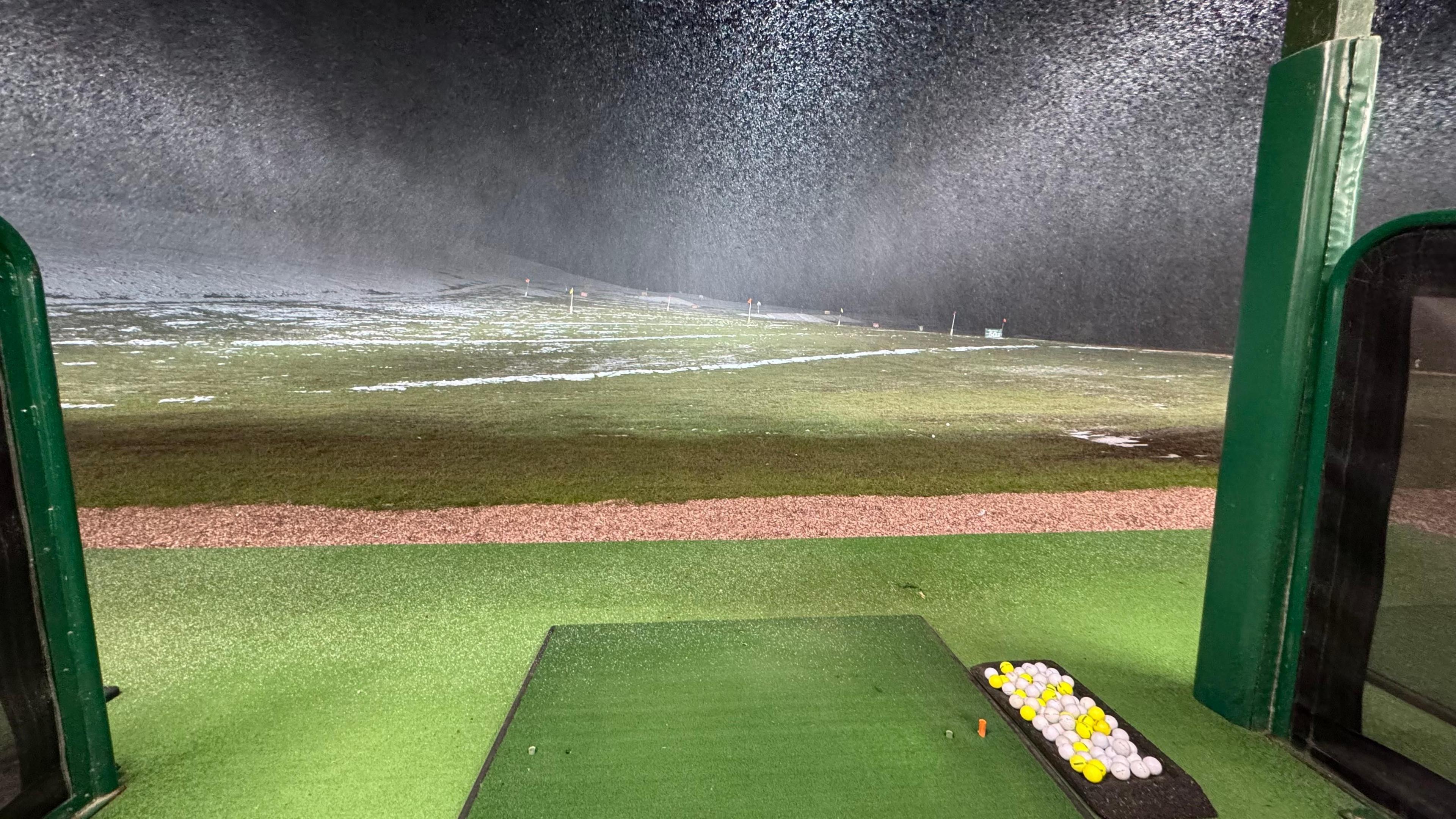 A shot at a golf driving range with snow starting to settle on a rising landscape in the dark. Snow is falling heavily. In the foreground, a tray of balls and mat to play them off are visible. 