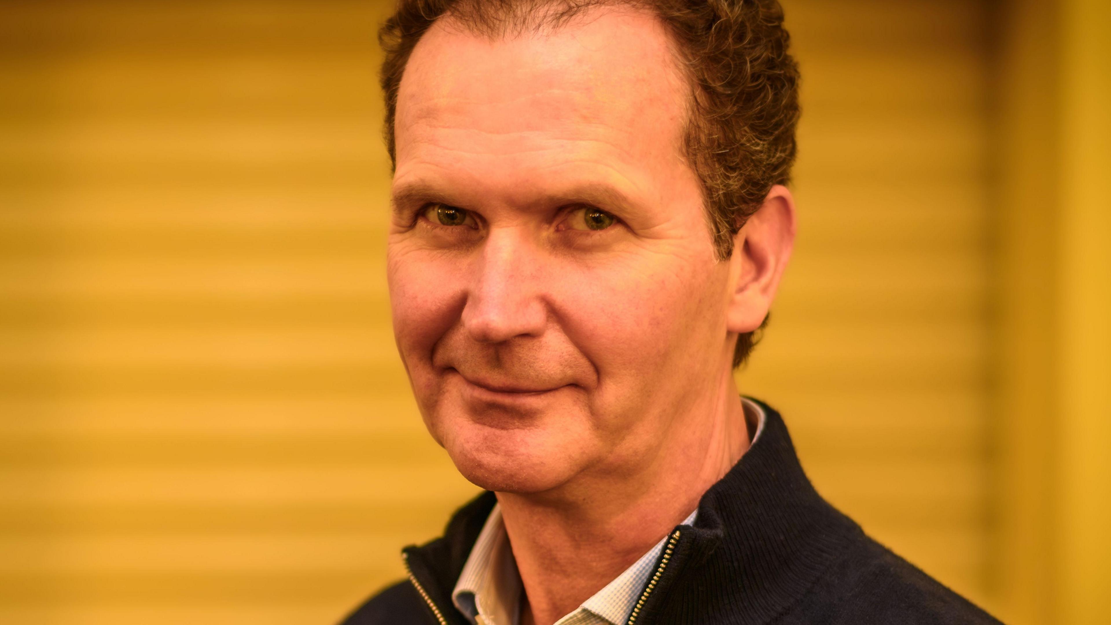 James Wilcox is wearing a white shirt with a black zipped jumper over it. He is looking at the camera sideways on and has dark, receding hair.