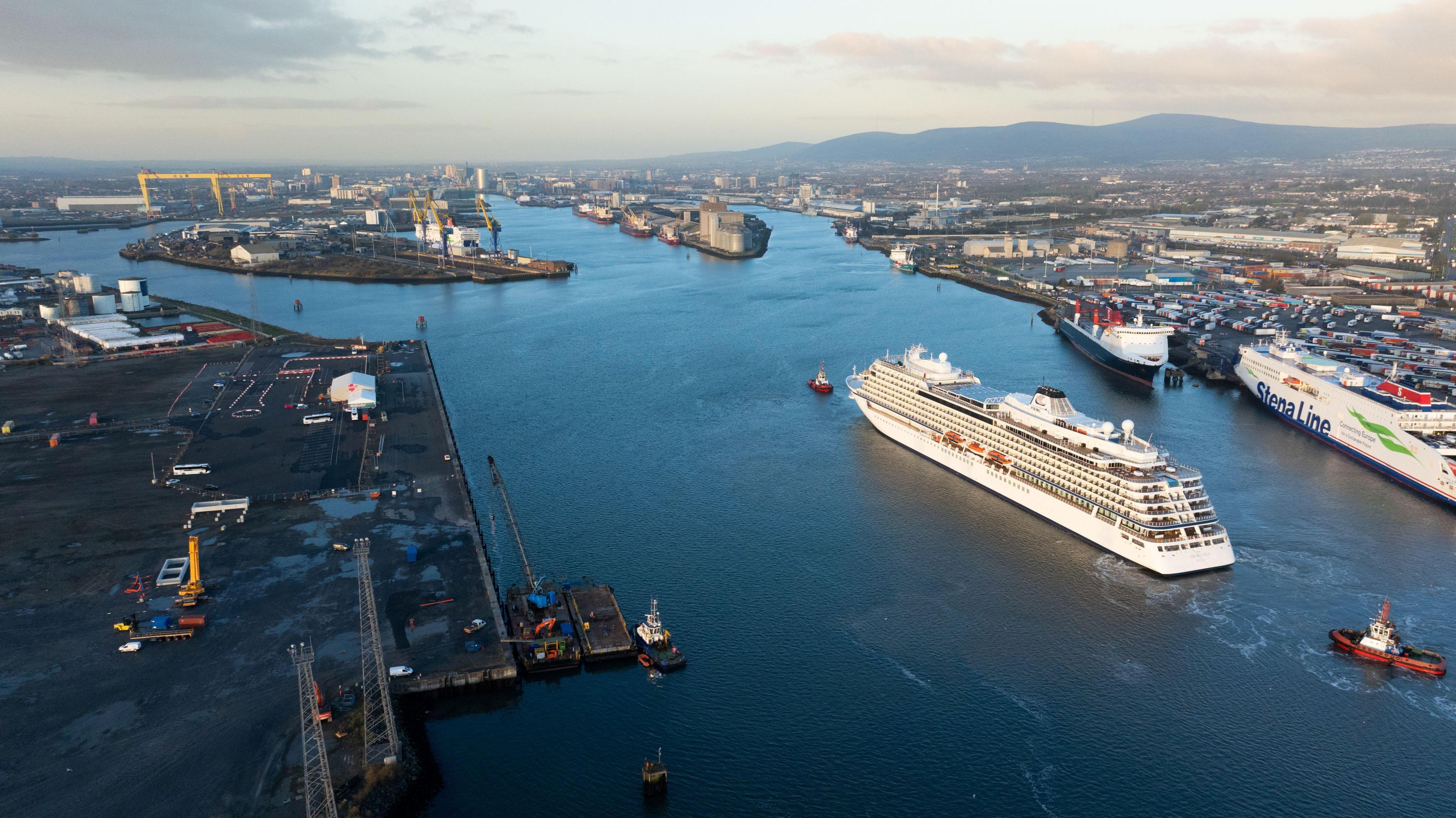 A shot from above of Belfast Harbour. There is water in the middle and land on either side. Two yellow cranes are visible in the distance. There is a cruise ship in the middle of the harbour and a Stena Line ferry on the right. There are also smaller boats.
