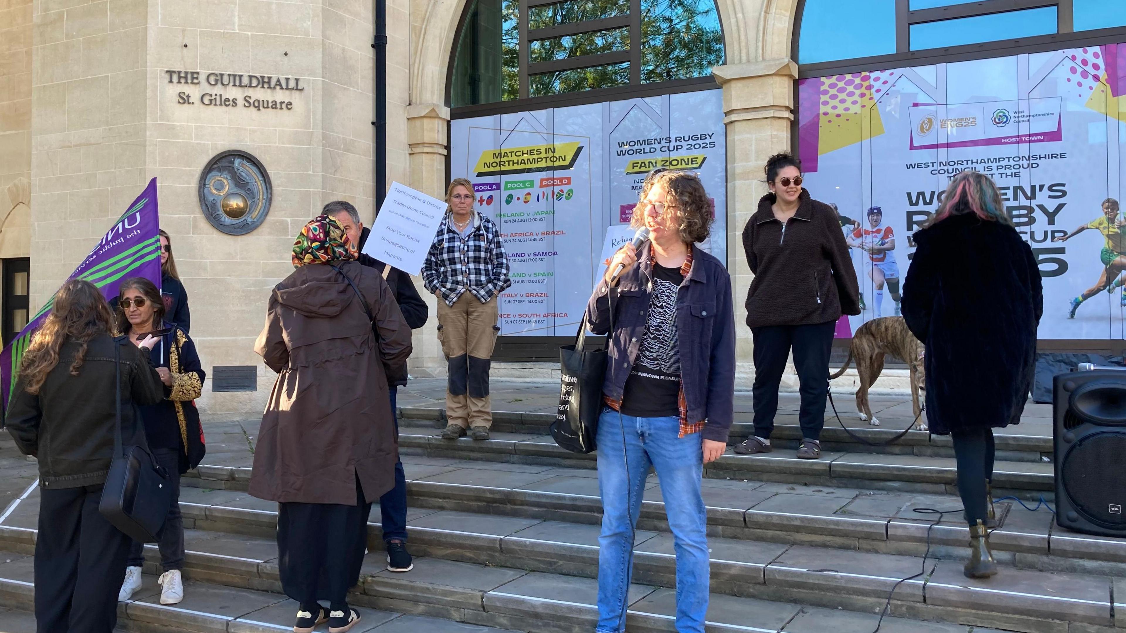 A small group of men and women on the steps of Northampton Guildhall. In the middle is a woman in blue jeans and a blue jacket over a black and white jumper with curling light brown hair holding a microphone up to her mouth as she addresses people. 