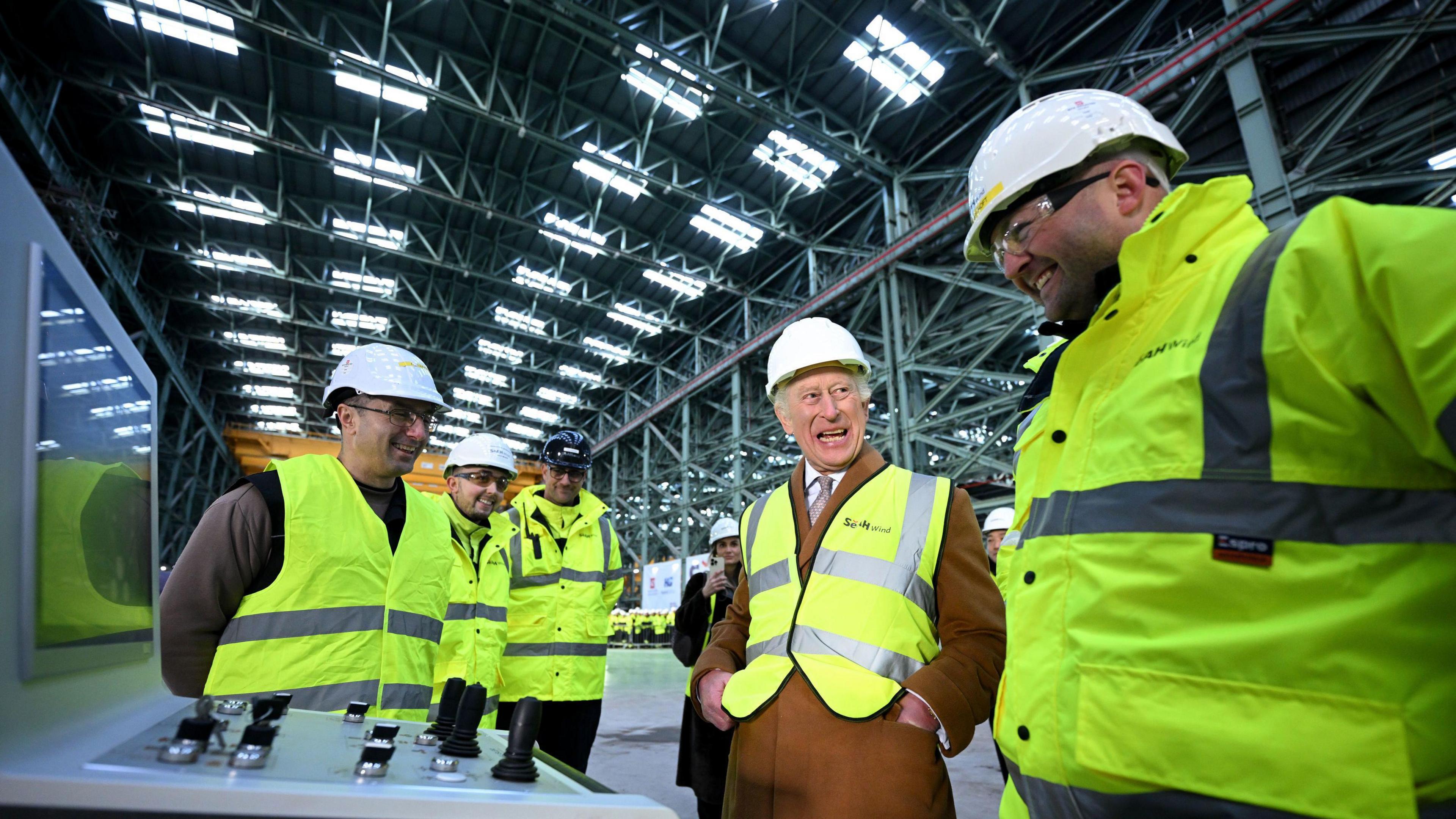 King Charles in a hard hat and hi-vis vest laughing with a man inside SeAH Wind's XXXL factory.